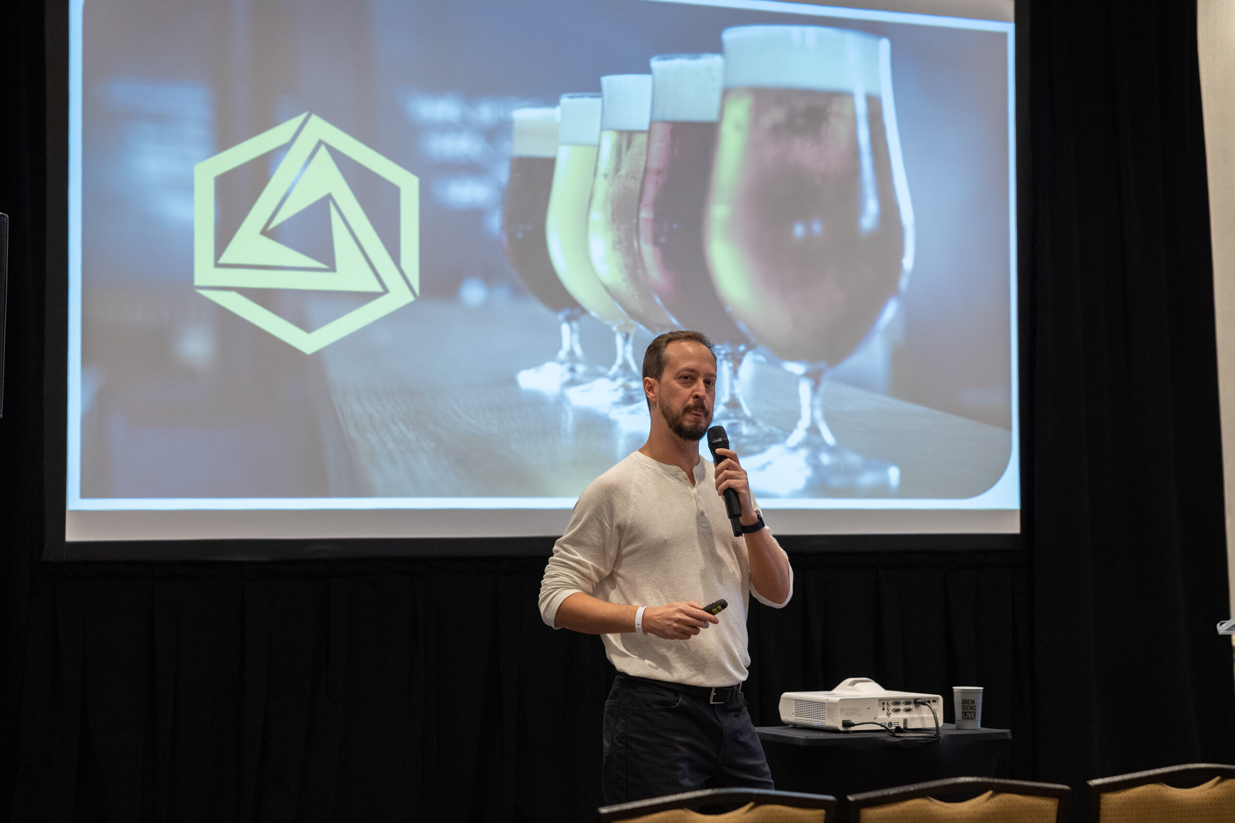 A man with a microphone stands before a screen showing the Abstrax logo and beer glasses.