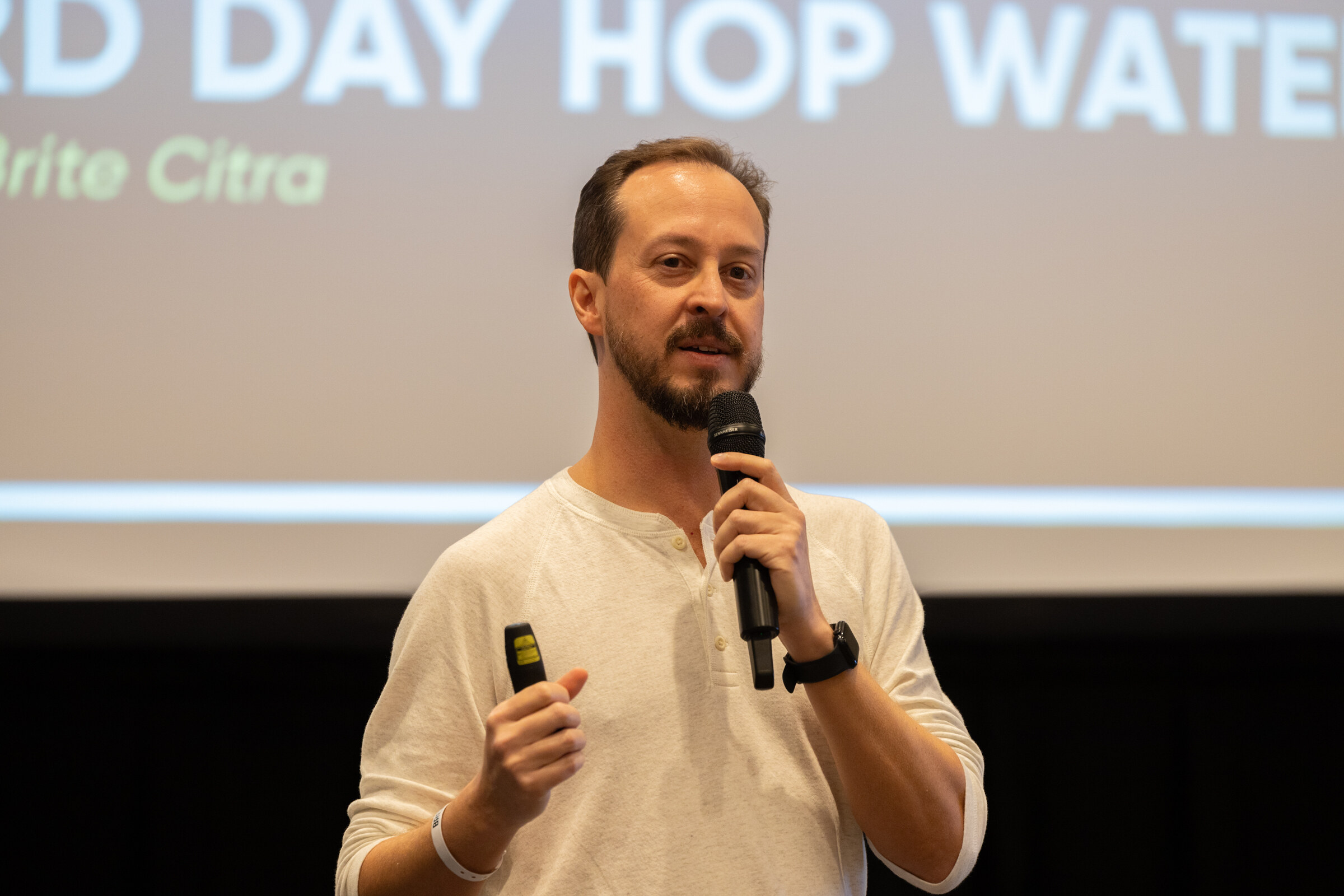 A bearded man holds a microphone and a clicker in front of a slide.