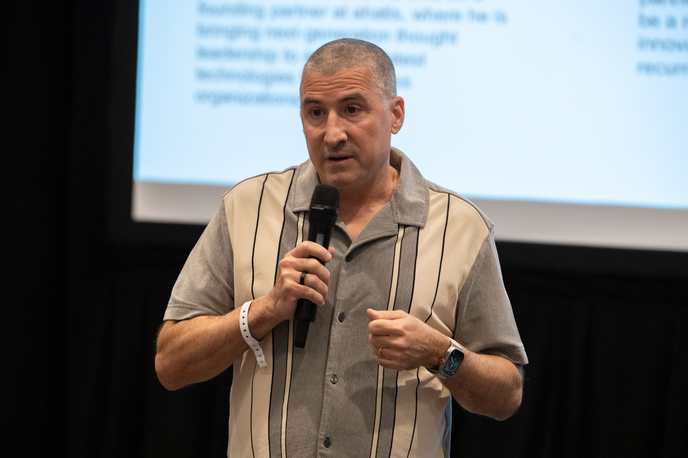A man in a gray striped shirt speaks into a microphone in front of a projected slide.