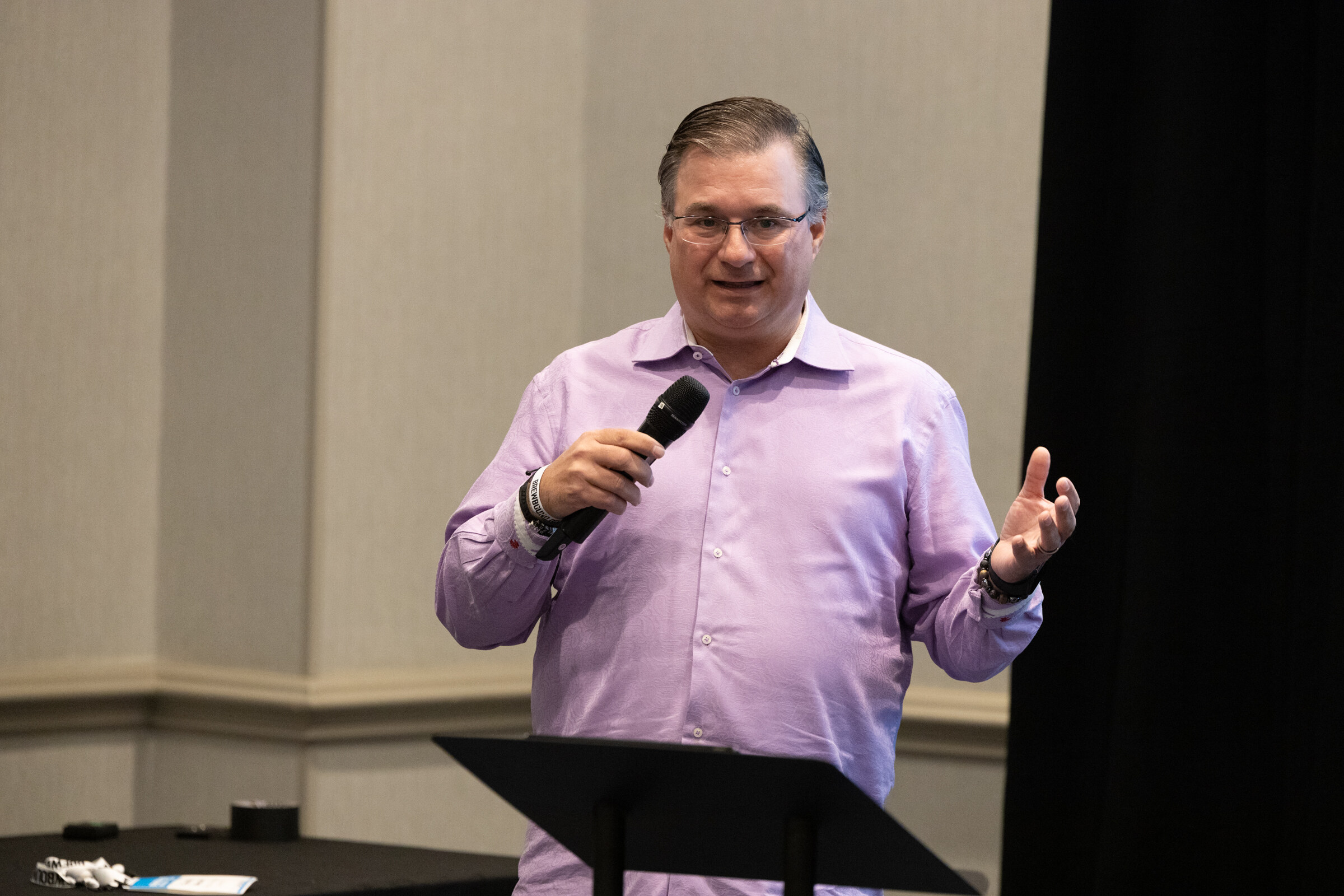A man in glasses and a lavender shirt speaks into a microphone at a podium.