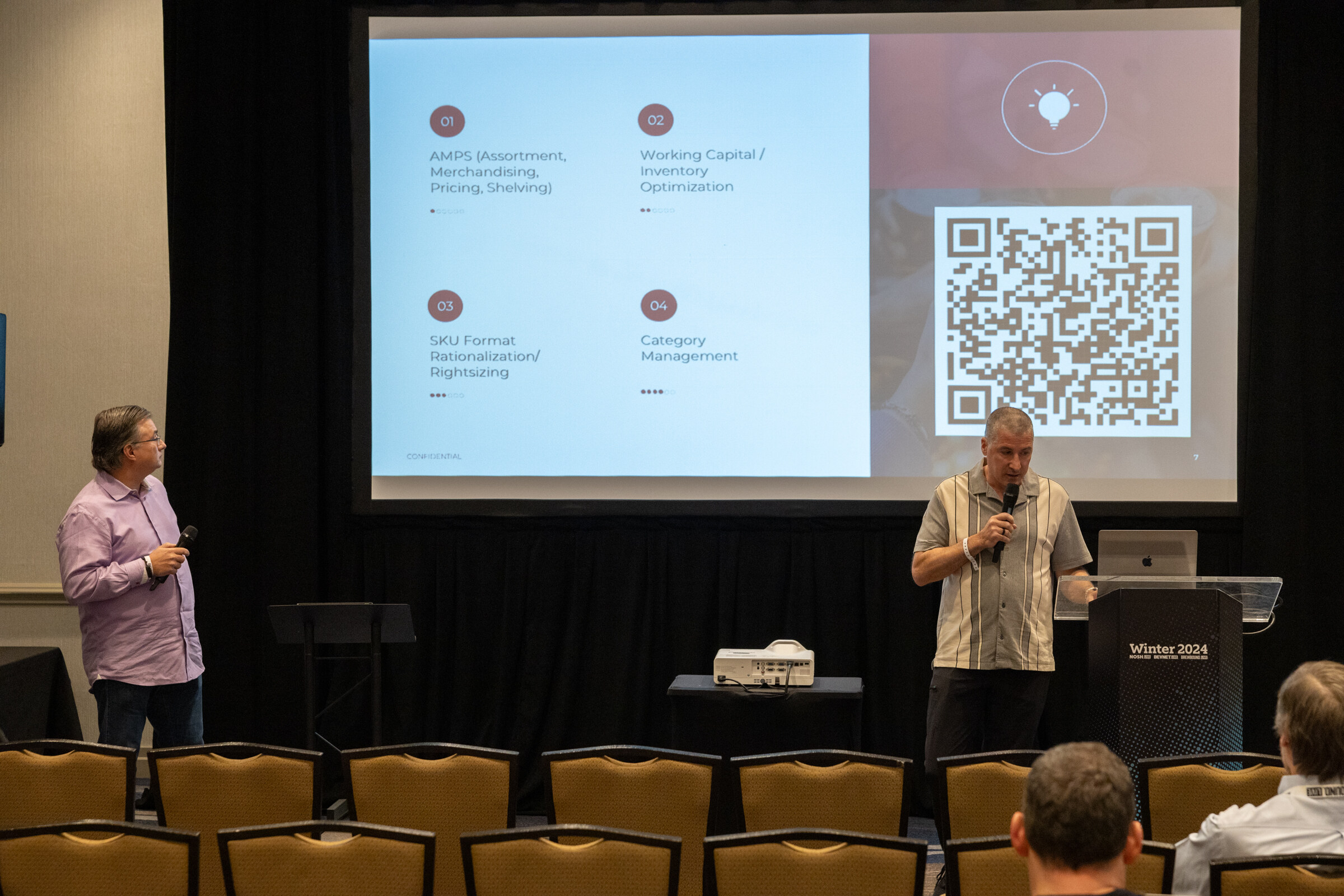 Two men stand in front of a screen with four numbered topics and a QR code.