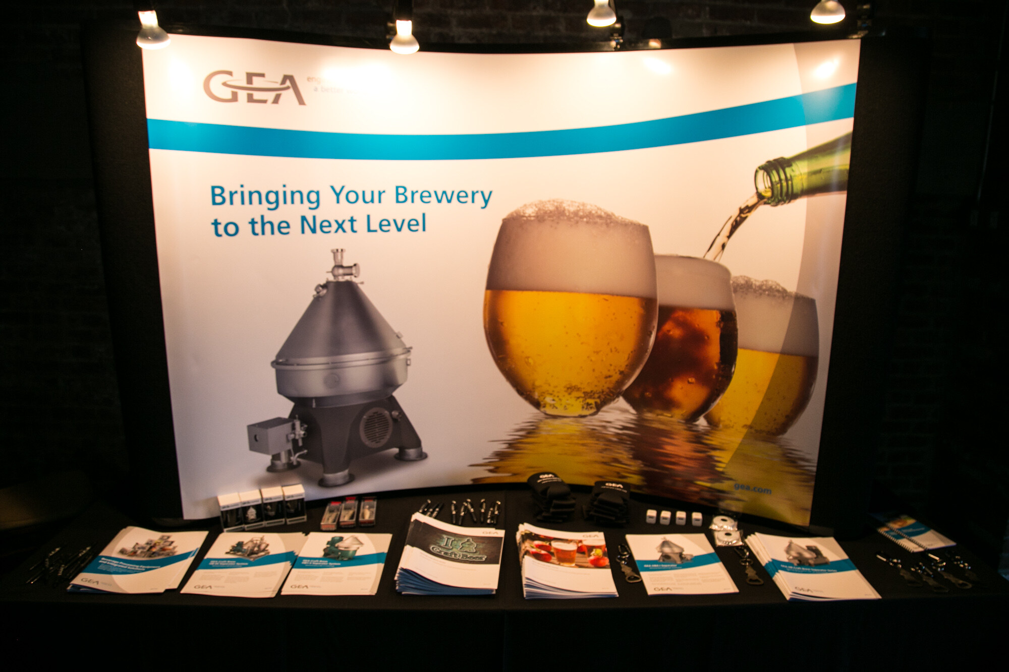 Exhibitor table with a GEA banner reading Bringing Your Brewery to the Next Level, and brochures on a black tablecloth.