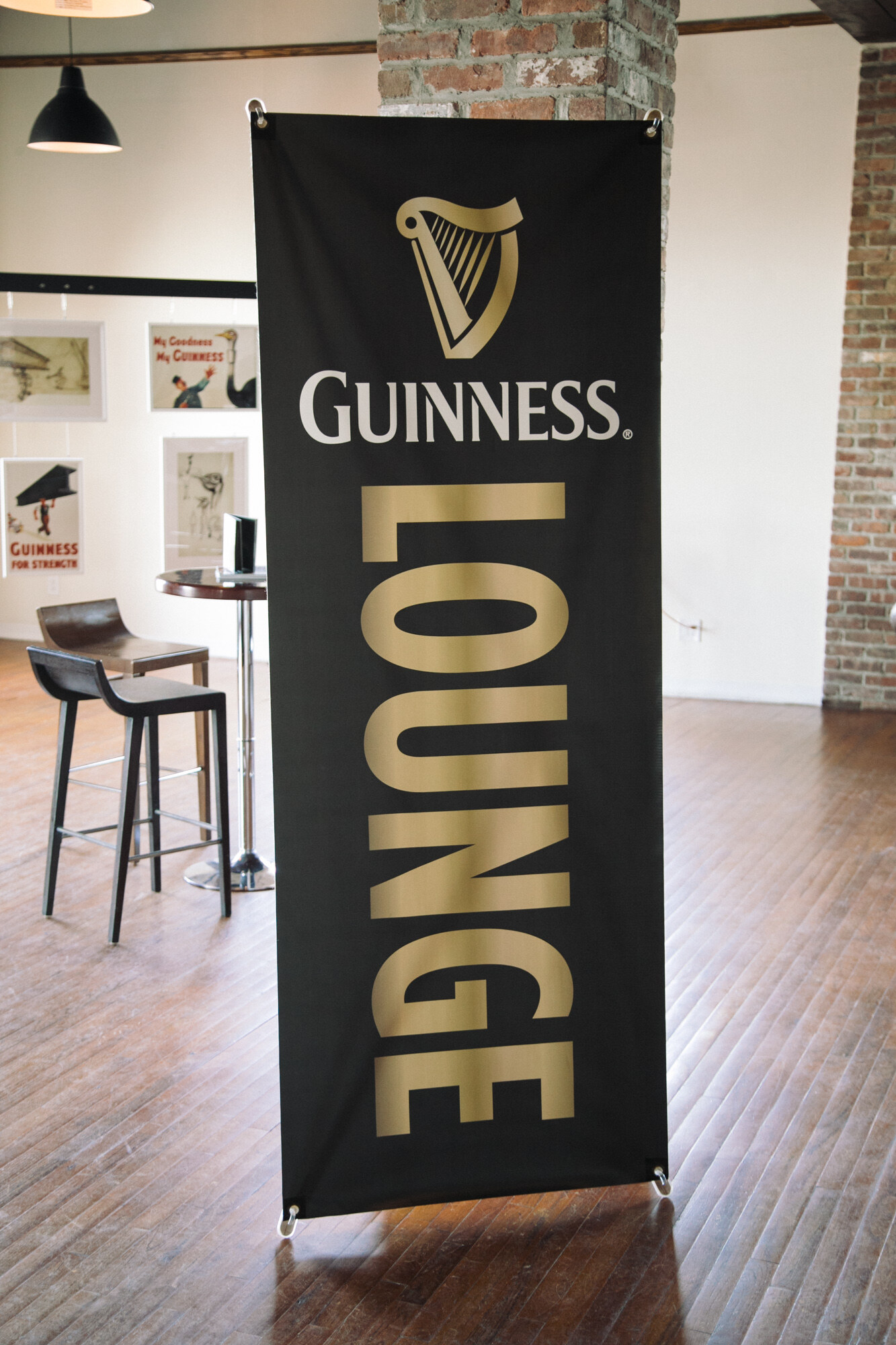 Tall black banner reading Guinness Lounge in gold letters on a wood floor, with bar stools and a brick wall behind.