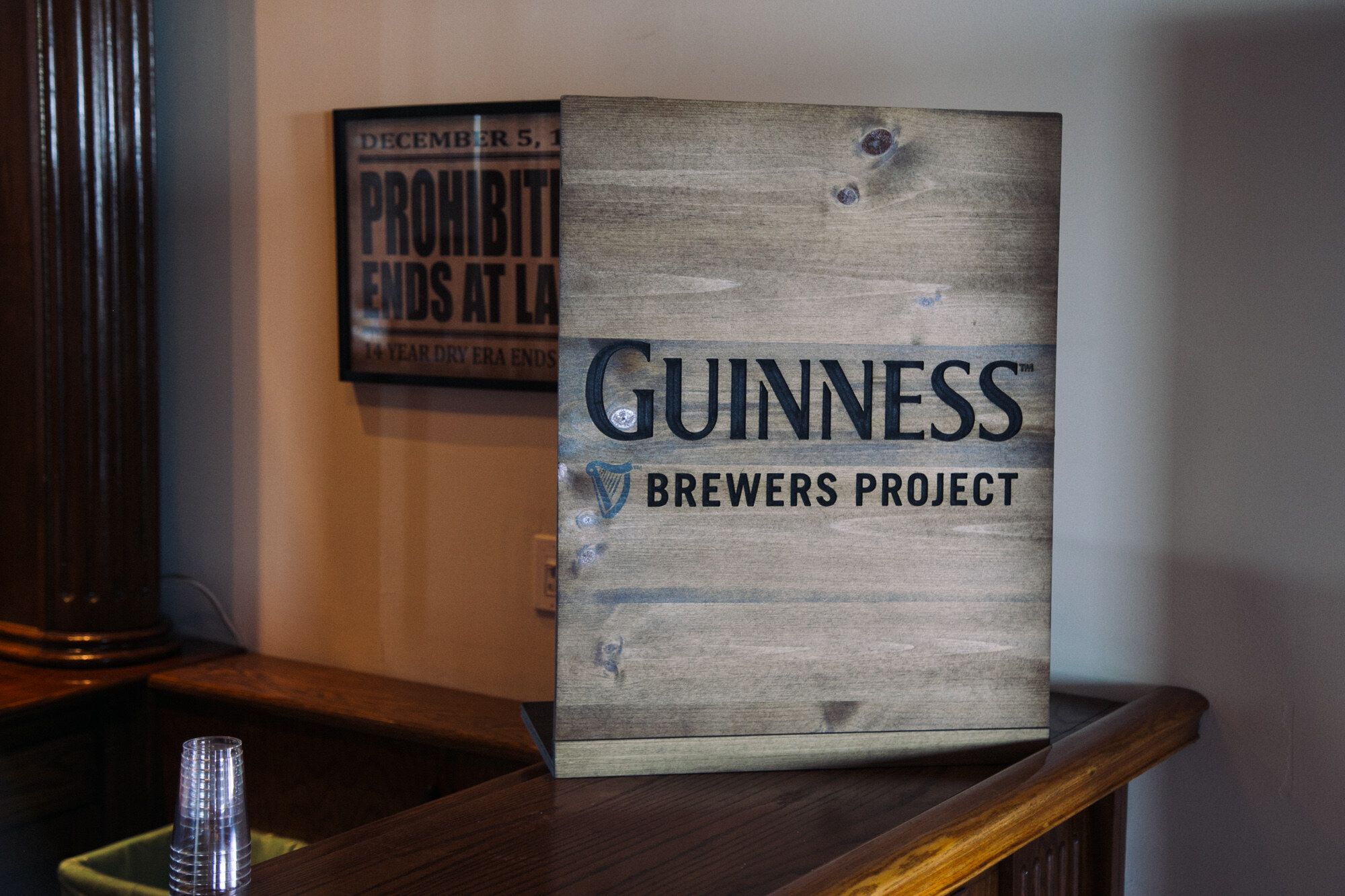 Wood-panel sign reading Guinness Brewers Project on a wooden bar with a plastic cup nearby.