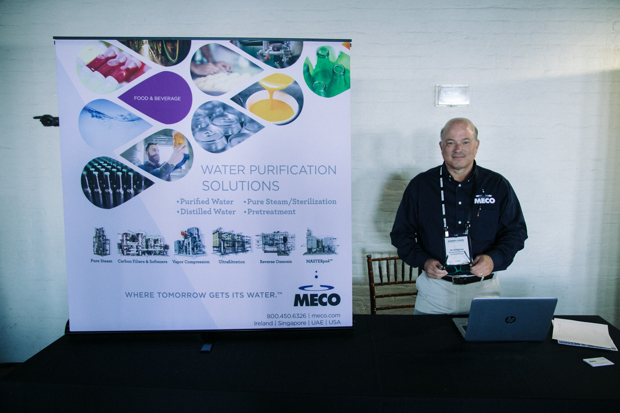 Man in a dark MECO shirt standing at a draped table with a laptop in front of a banner reading Water Purification Solutions.