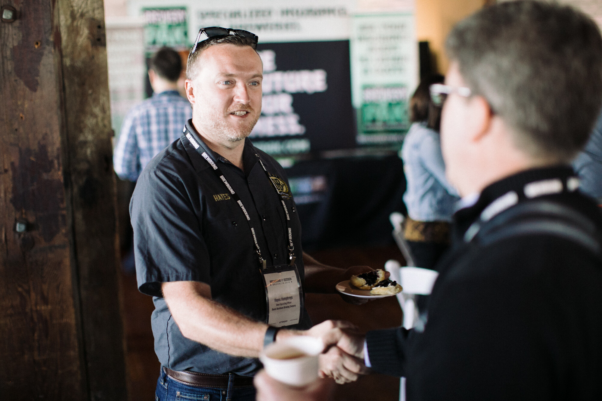 Man in a dark work shirt with a lanyard shaking hands with another attendee, holding a plate with a pastry.