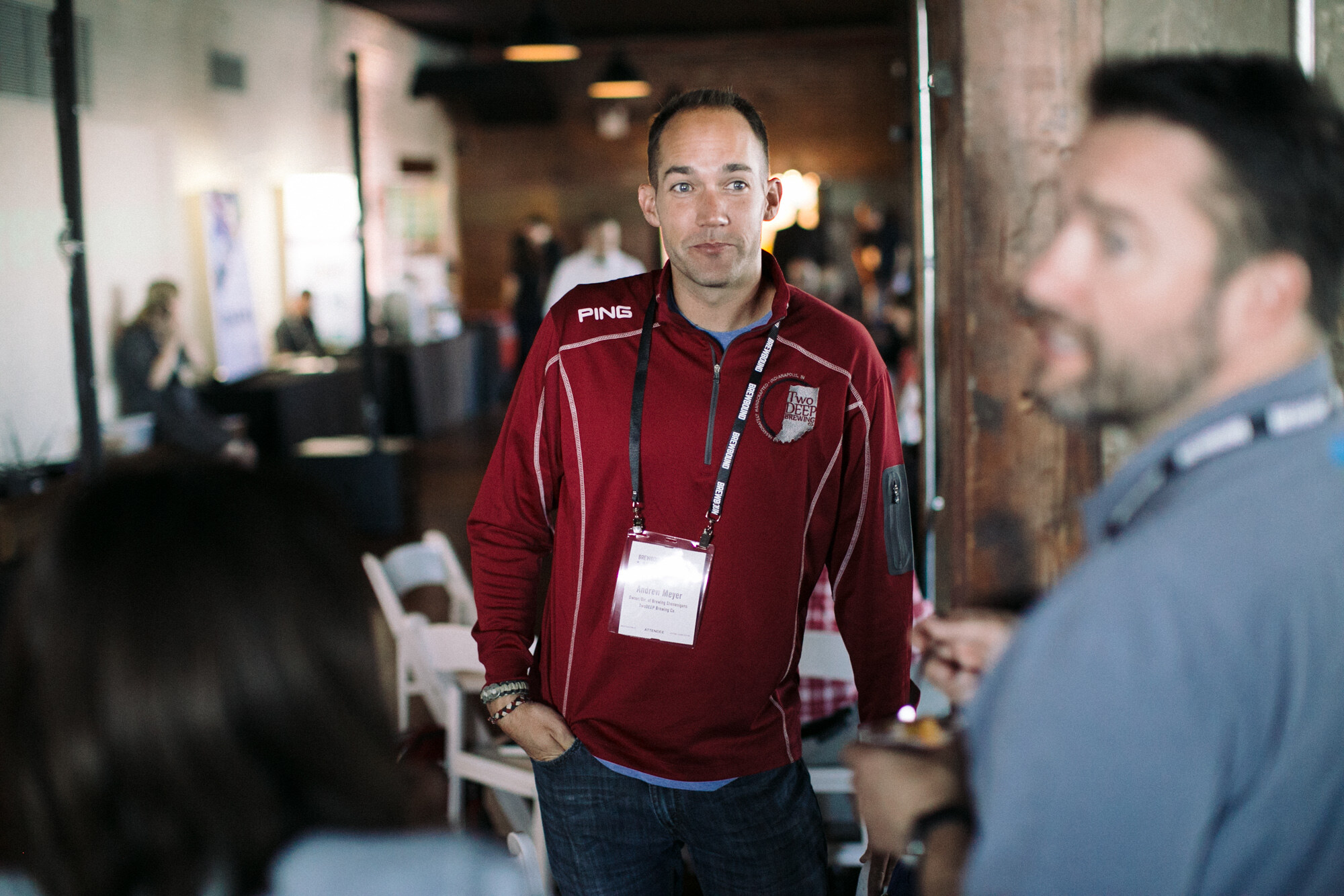 Man in a red quarter-zip pullover with a lanyard and name badge standing in a brick room with other attendees.