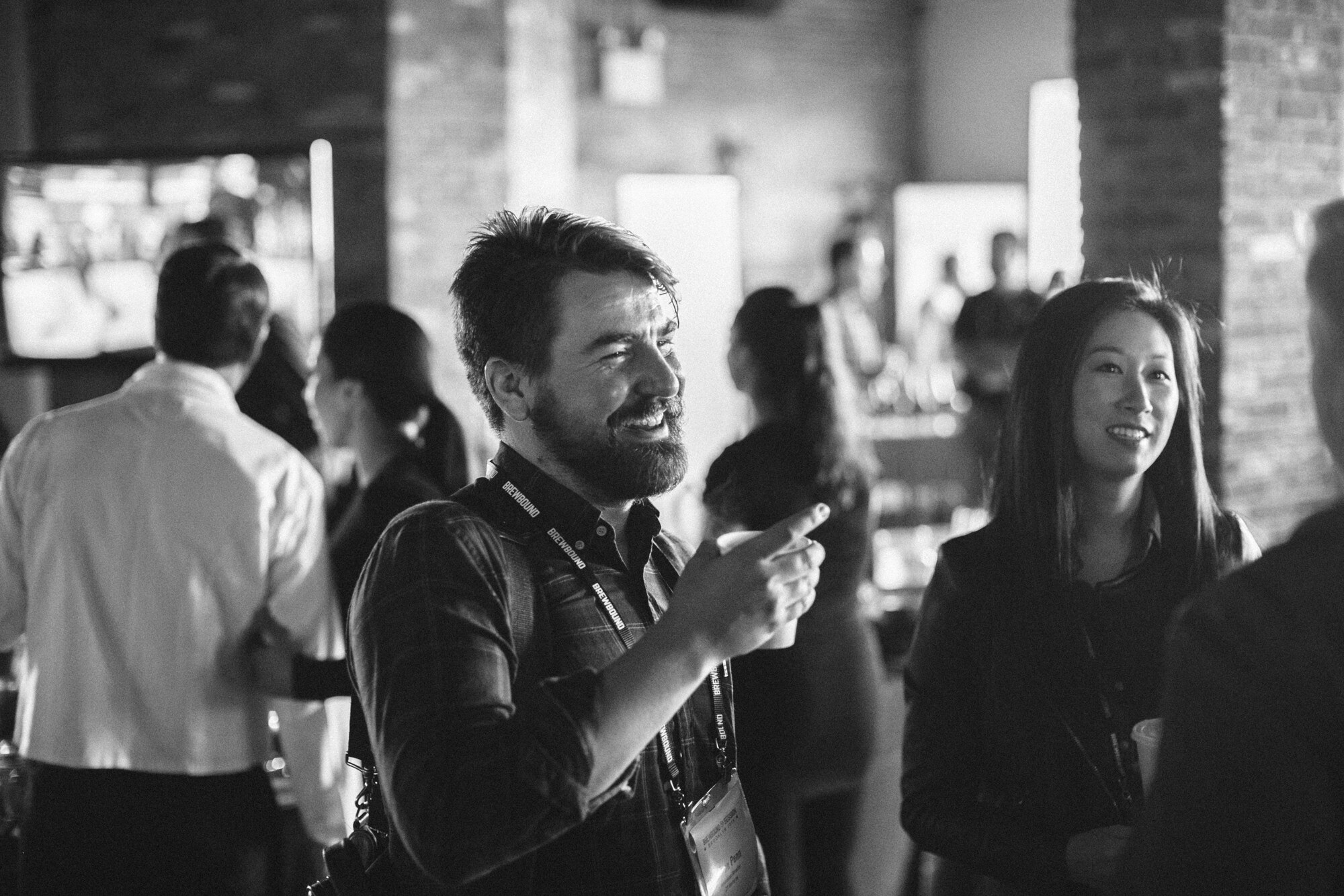 Black-and-white photo of a bearded man gesturing with a cup and a smiling woman in a crowded brick room.