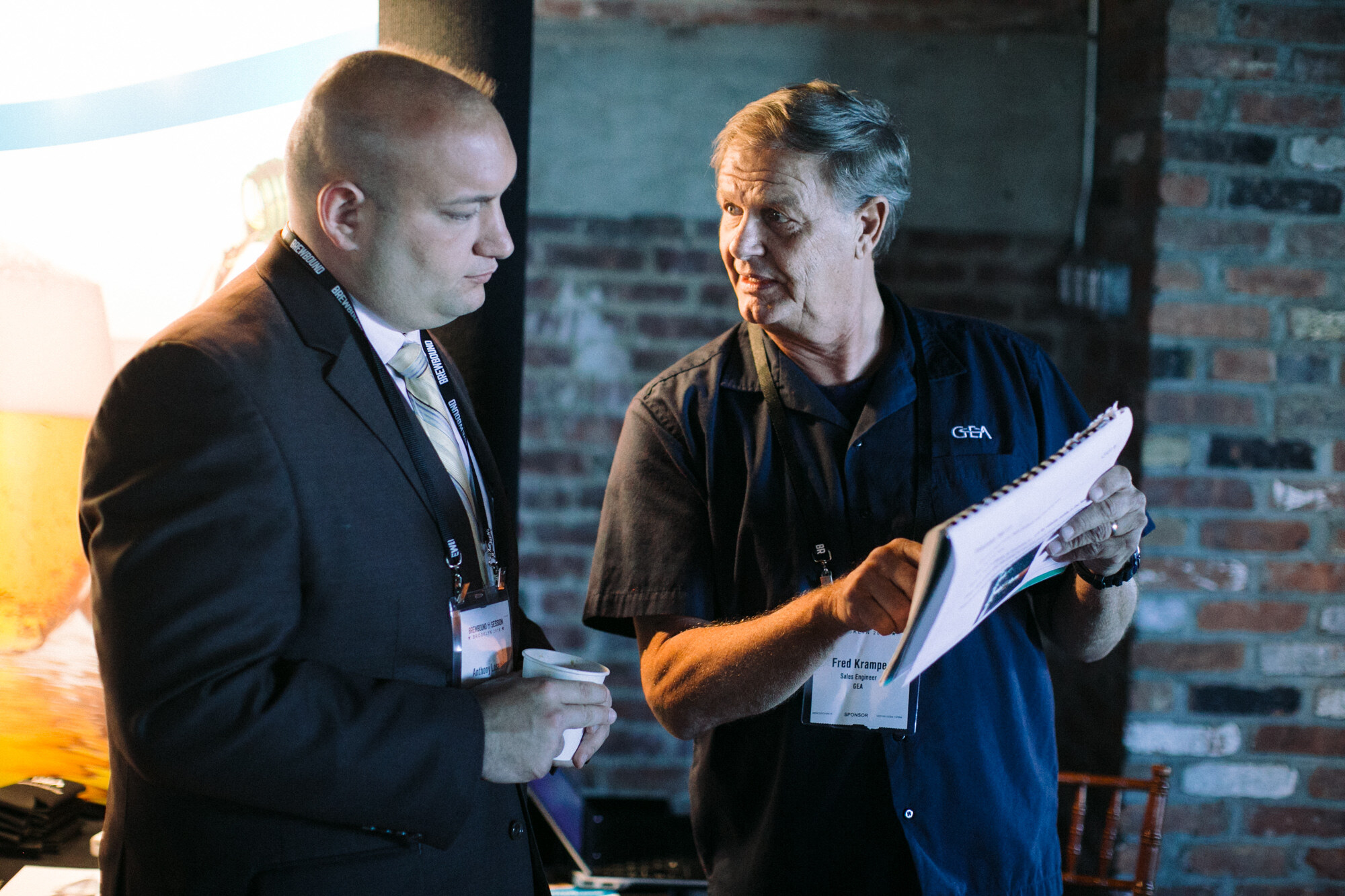 Man in a dark suit looking down and a man in a GEA work shirt pointing at a notebook, in front of a brick wall.