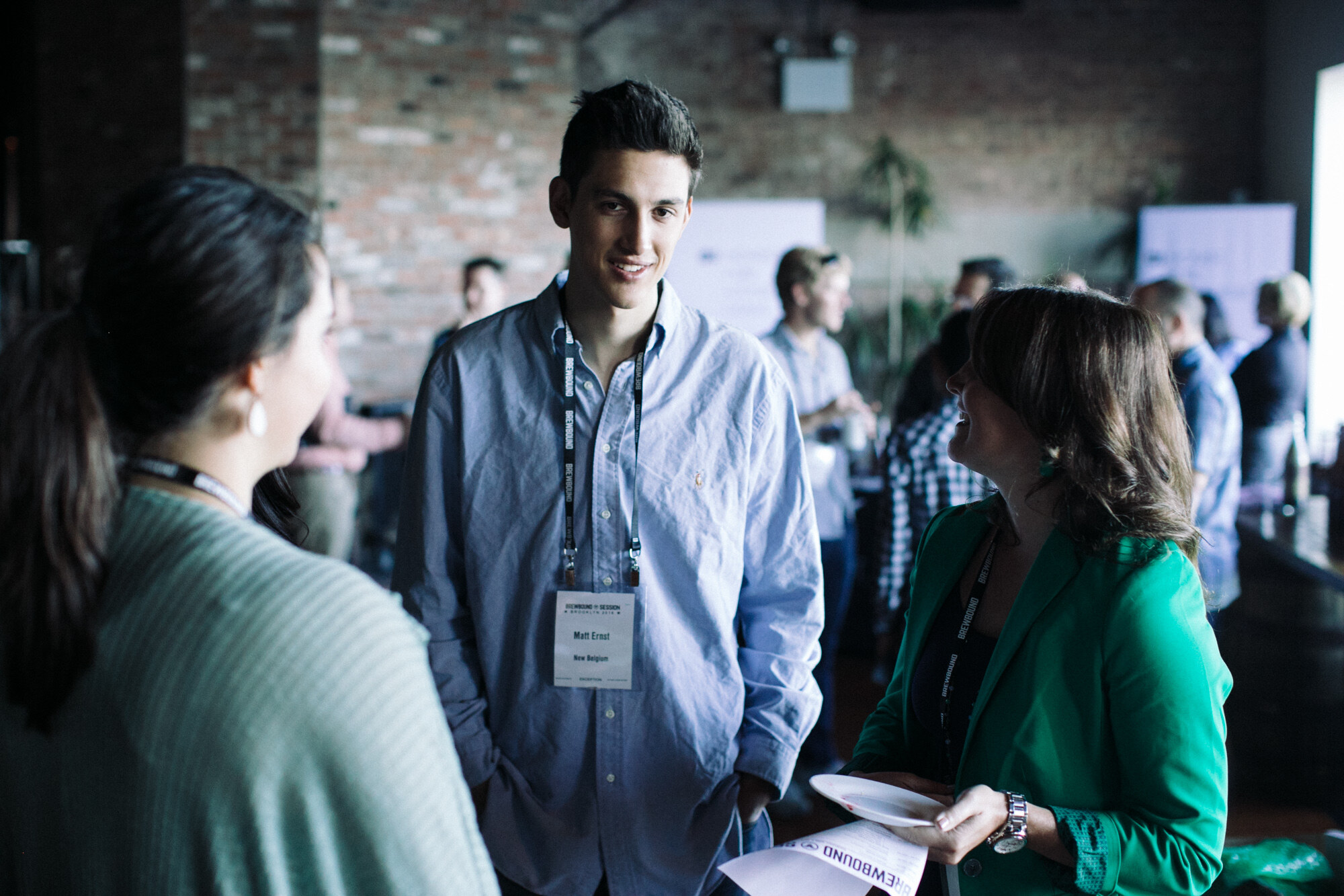 Young man in a light blue shirt with a lanyard listening to two women in front of a brick wall.