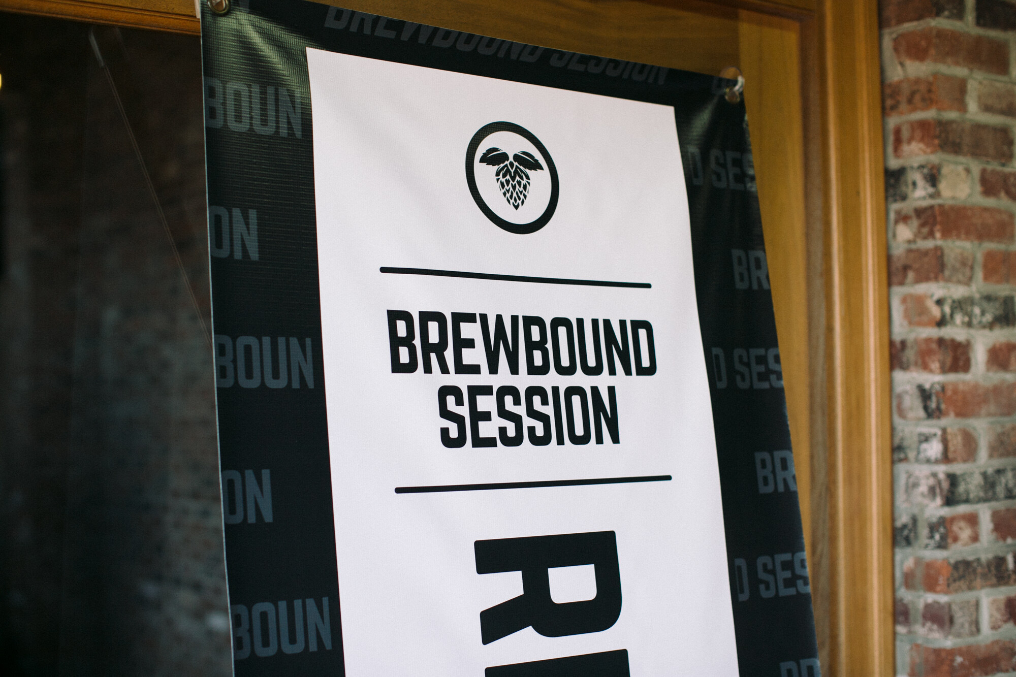 White and black banner reading Brewbound Session with a hop logo, hung in a wooden door frame beside a brick wall.