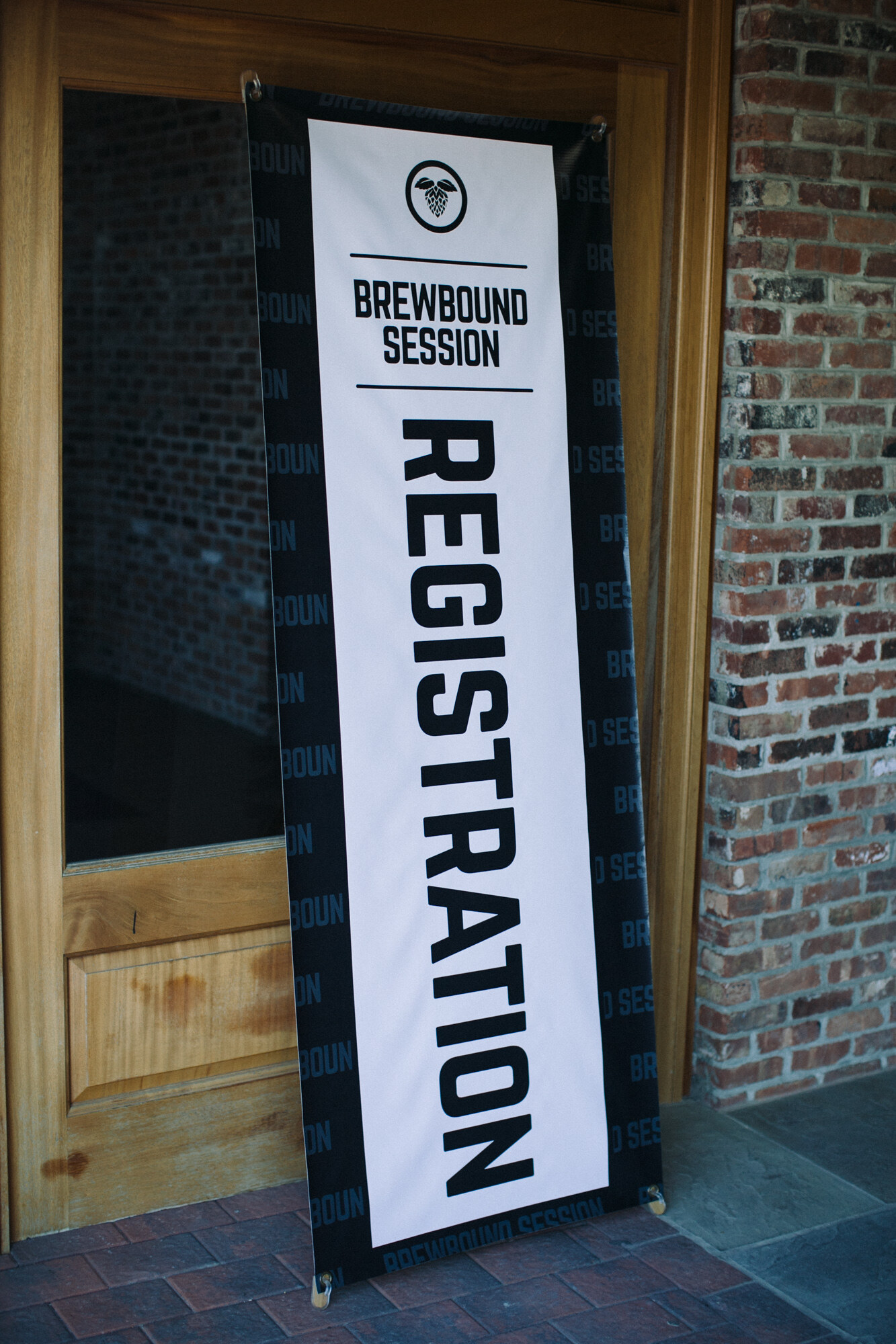 Tall black and white banner reading Brewbound Session Registration standing in a wooden doorway beside a brick wall.