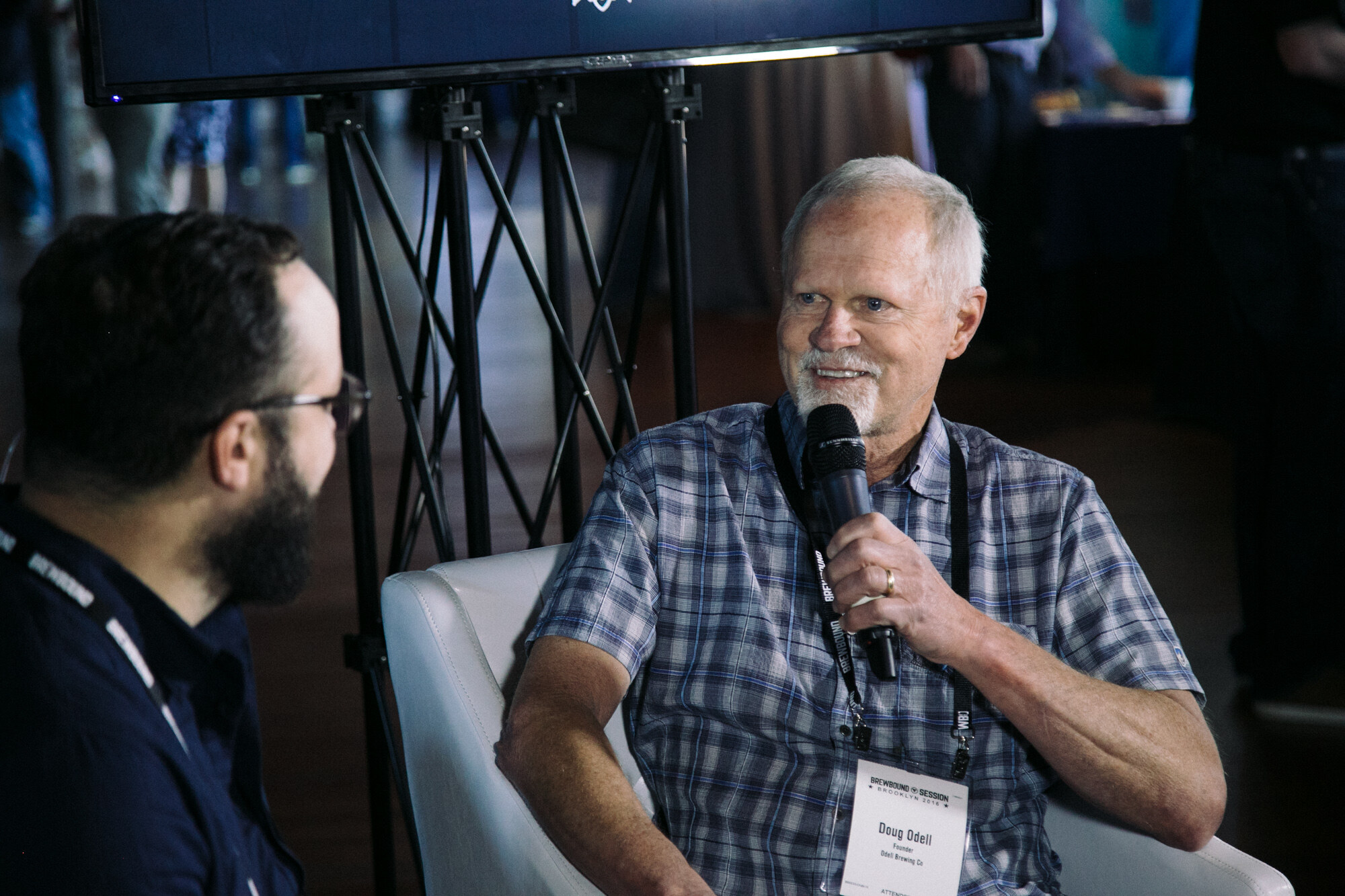 Gray-haired man in a plaid shirt holding a microphone, seated in a white armchair, with a bearded interviewer in the foreground.