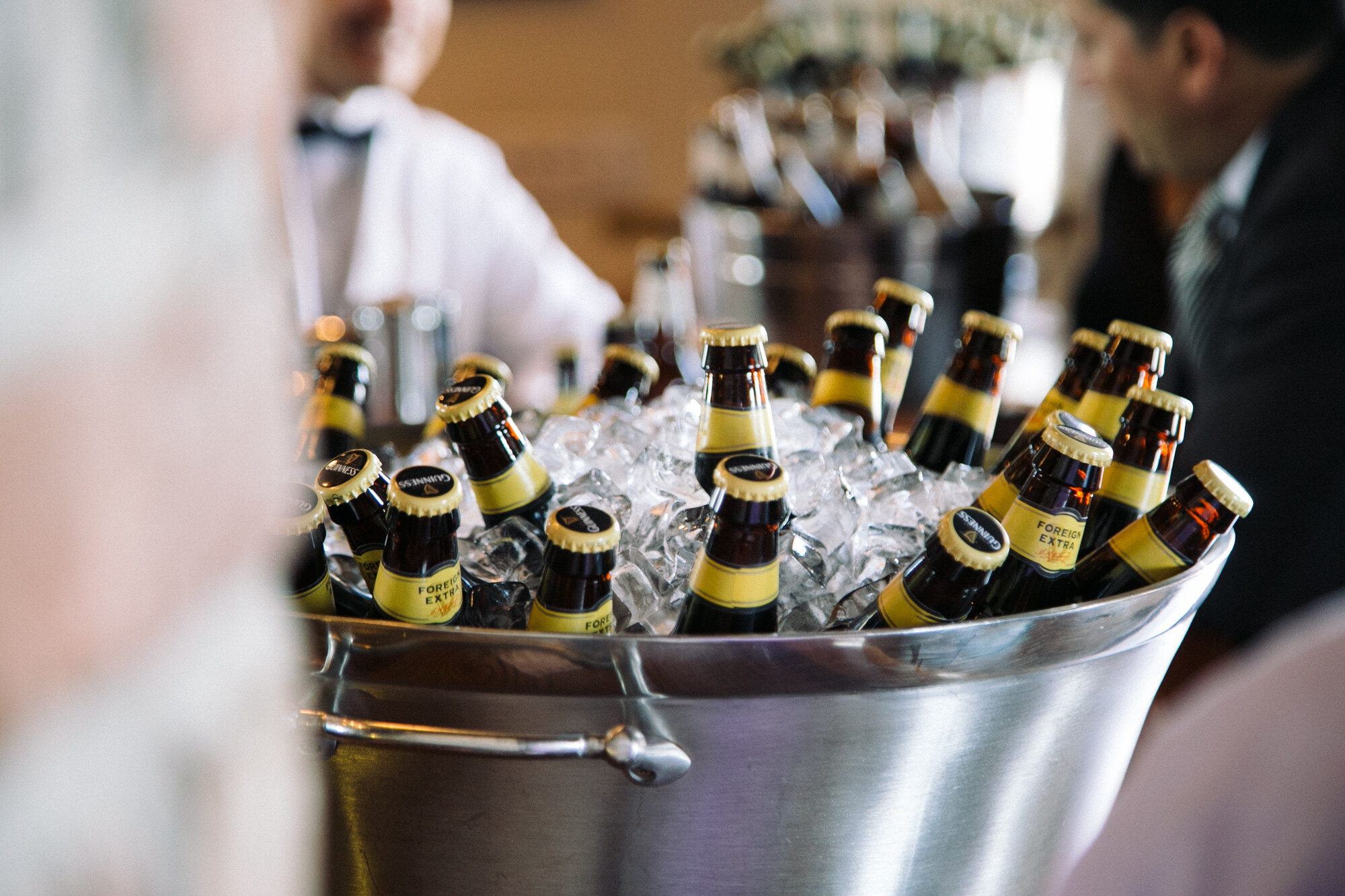 Silver bucket of ice filled with brown bottles with yellow-and-black labels, with a bartender in a white jacket and bow tie behind.