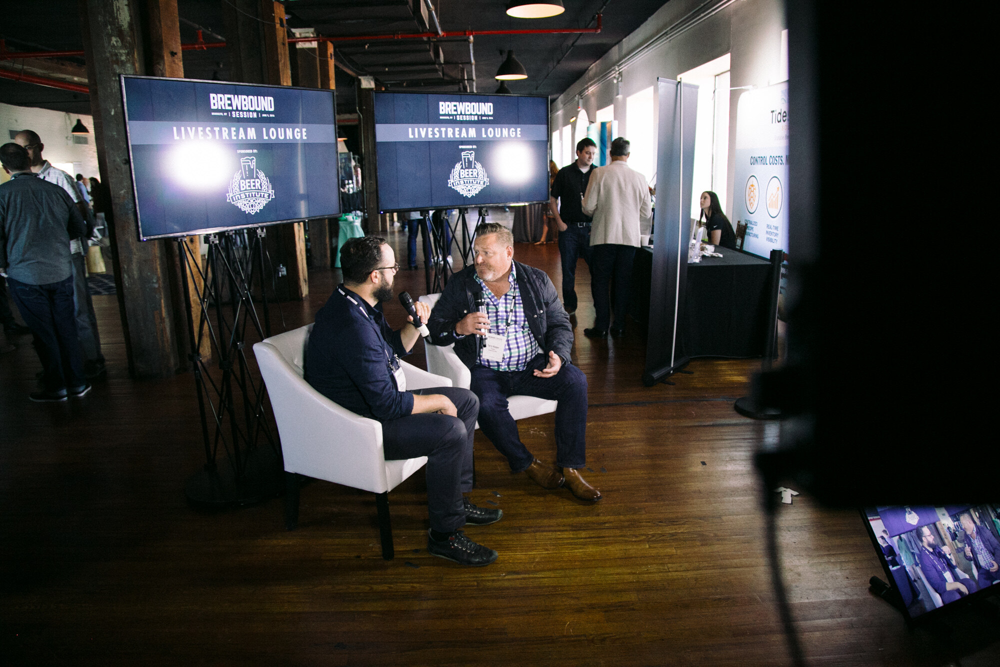Wide view of two people seated in white armchairs on a wood floor between two screens reading Livestream Lounge, with exhibitors' tables behind.
