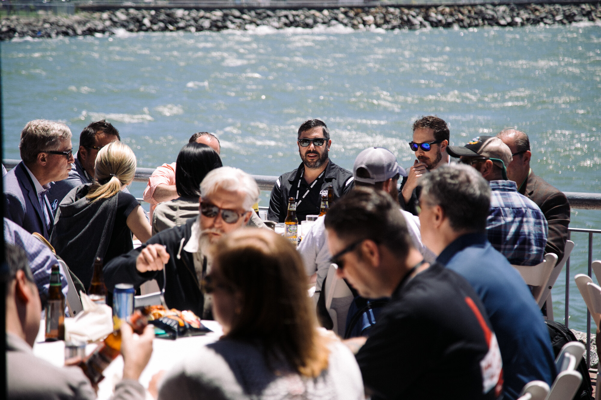 Groups of people seated at white tables with beer bottles on a terrace beside choppy water.