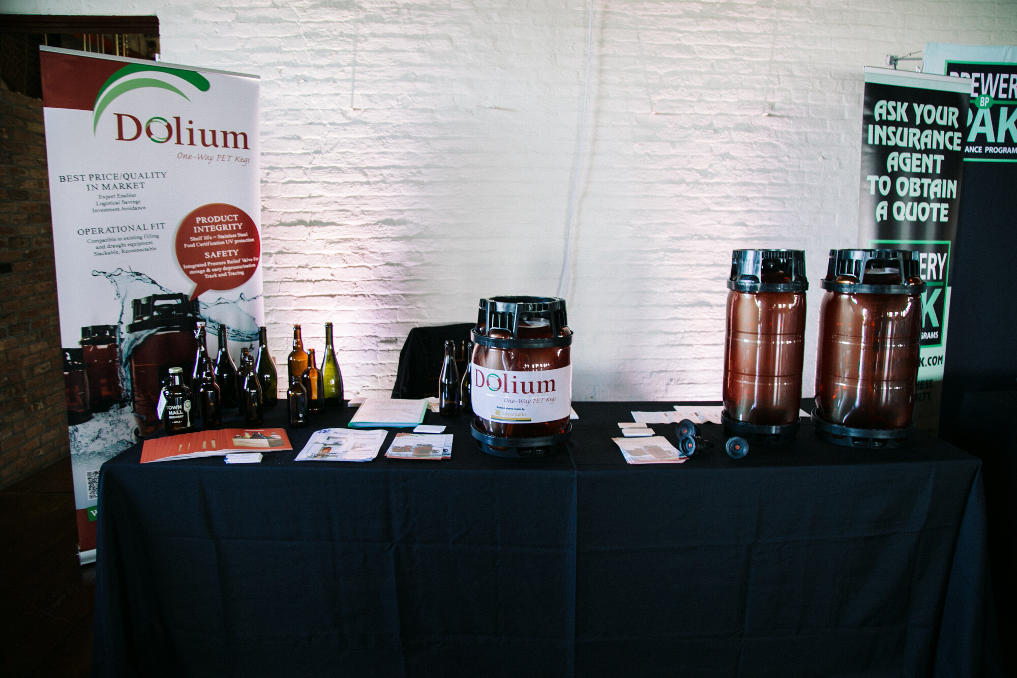Black-draped table with several brown plastic kegs and a Dolium banner, plus a Brewery Pak insurance banner at right.
