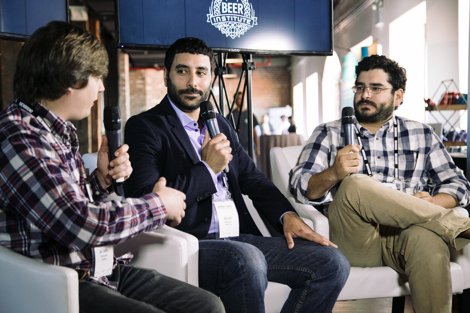 Three men holding microphones seated in white armchairs beneath a Beer Institute logo screen in a bright, windowed room.