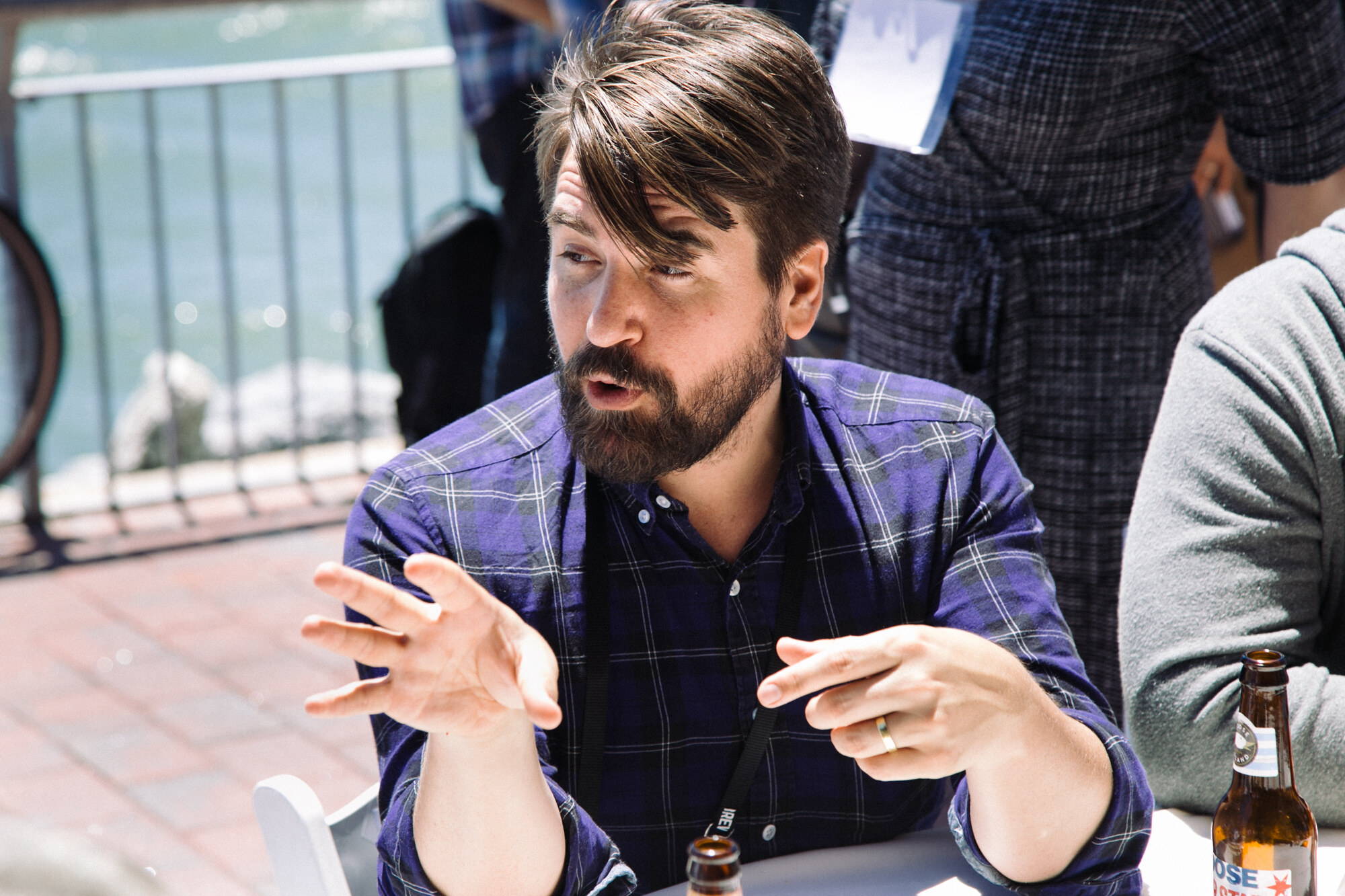 Bearded man in a purple plaid shirt gesturing with both hands, a beer bottle at the lower right, with a railing and water behind.