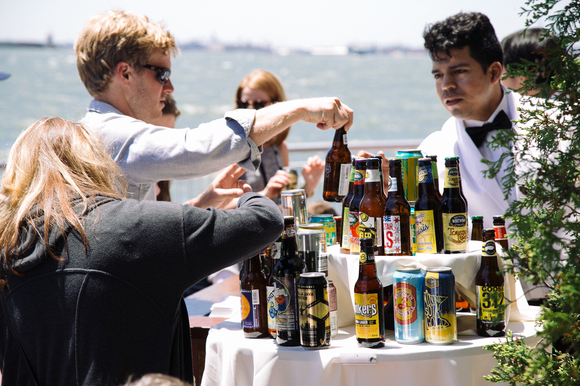 Table with white cloth covered in assorted beer bottles and cans, with a bartender in a bow tie and attendees reaching for bottles, water behind.