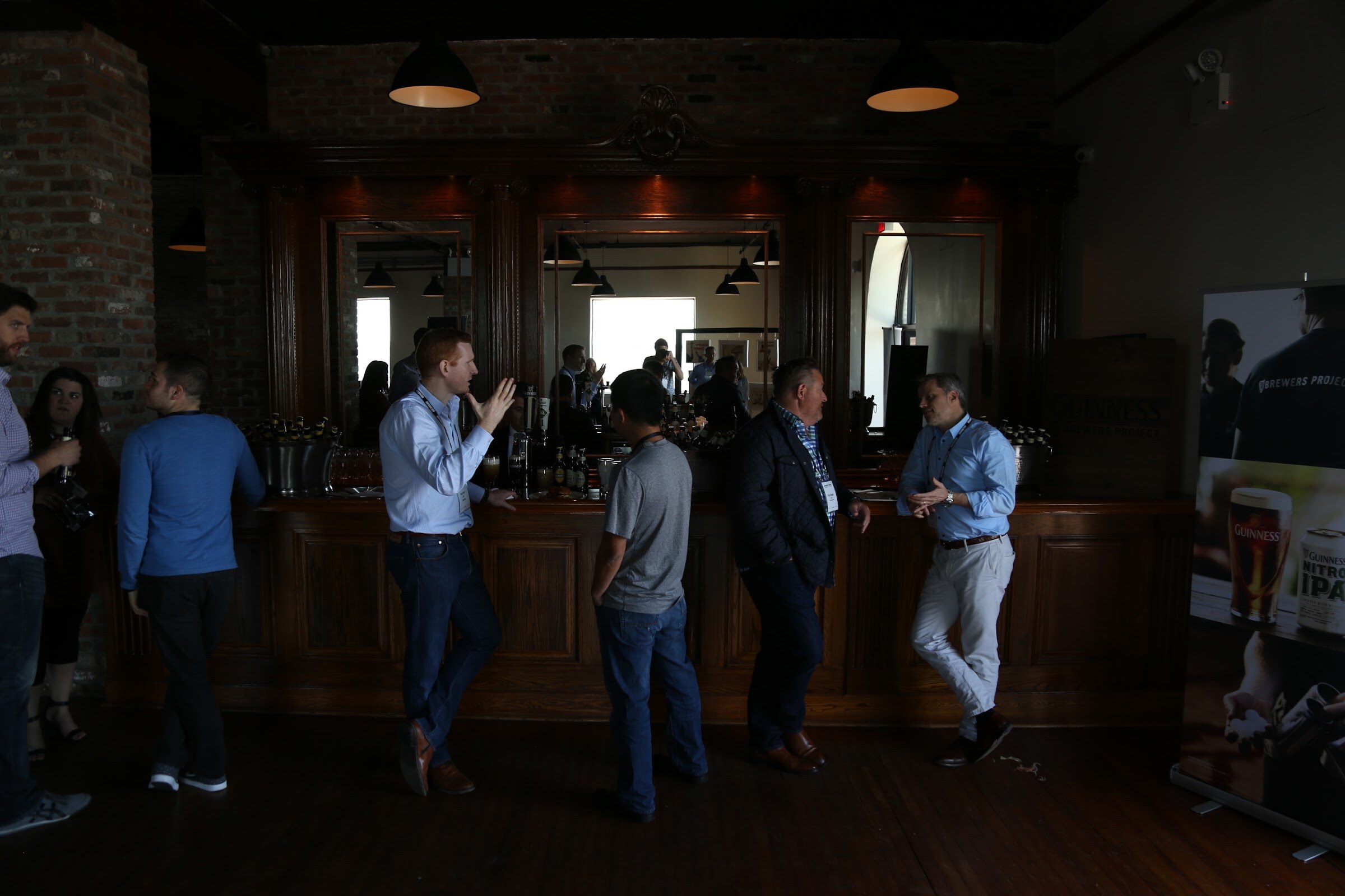Several attendees standing along a long wooden bar in a dim brick room, with a Guinness banner at the right.