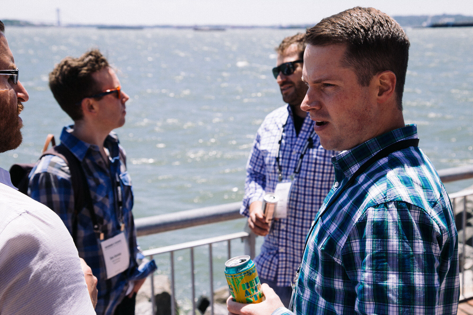 Two men in plaid shirts holding cans, talking by a metal railing with choppy water behind.