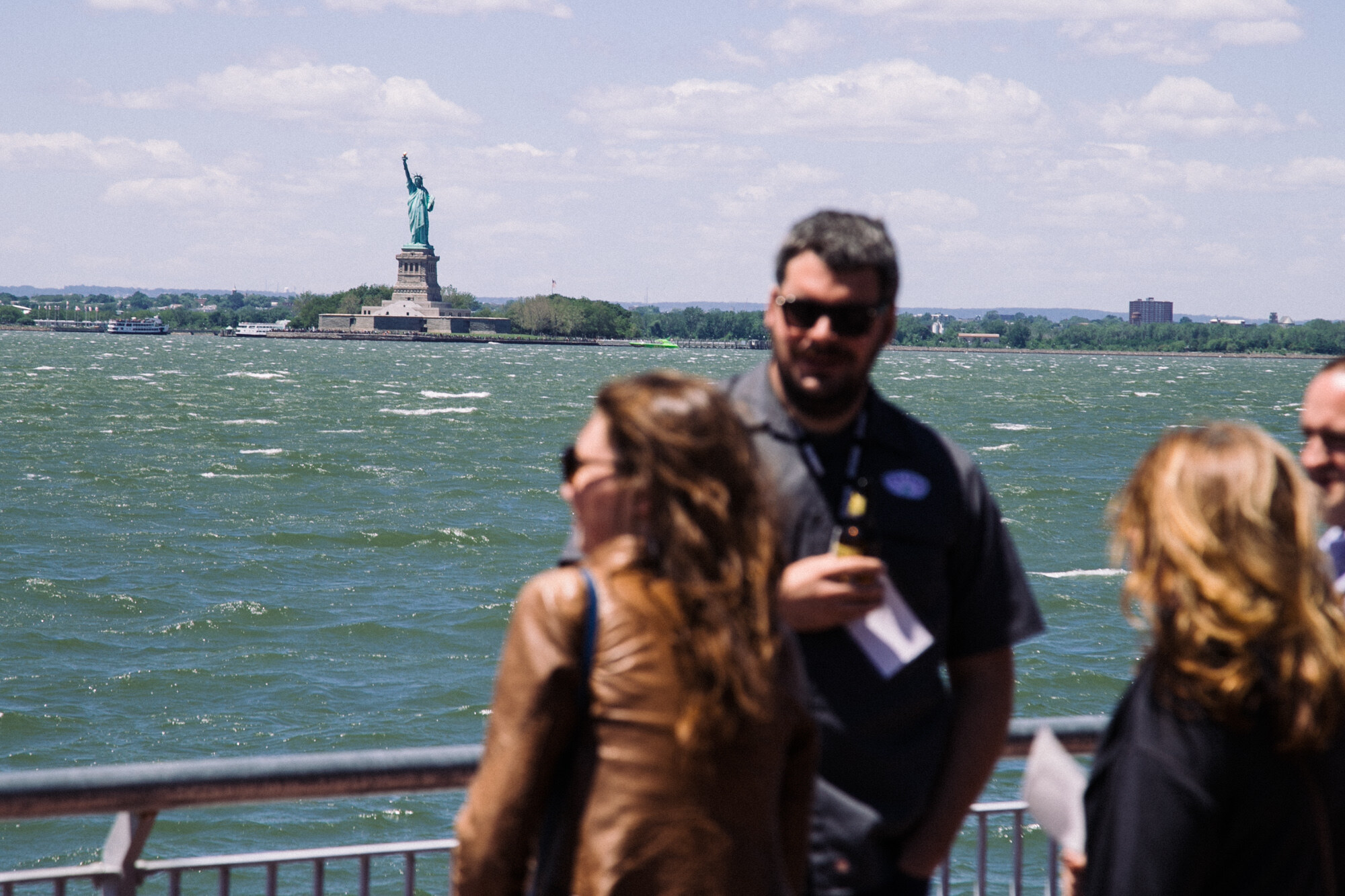 People standing at a railing in the foreground, blurred, with the Statue of Liberty on its island across green water.
