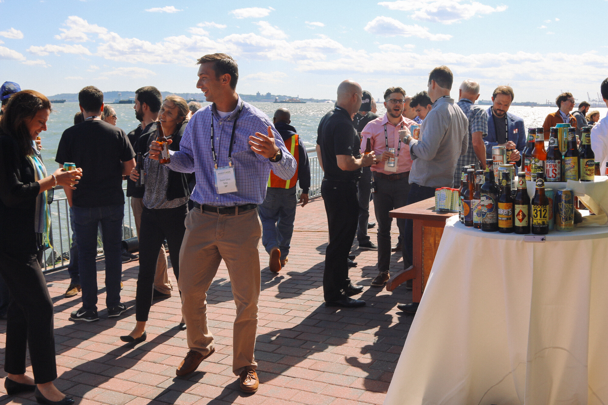 Crowd of attendees with lanyards standing on a brick-paved pier beside a table of beers, with water and sky behind.