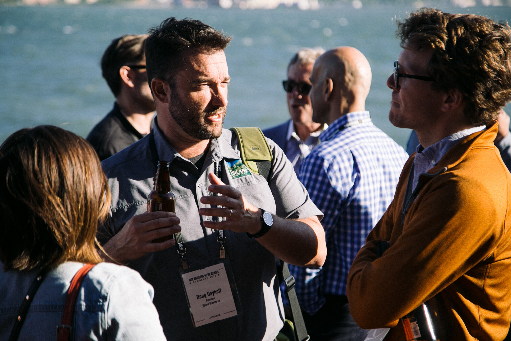 Bearded man in a gray shirt with a lanyard holding a beer and gesturing while talking with two other attendees by the water.
