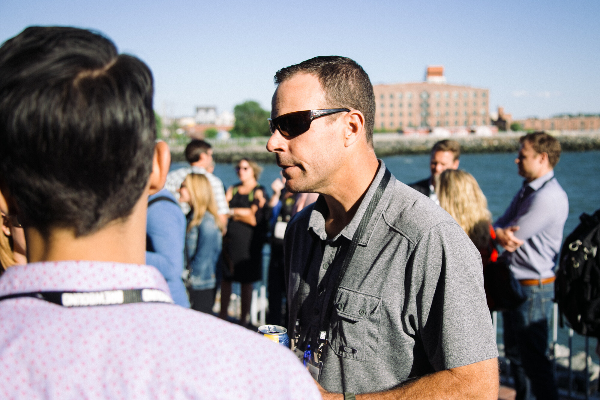 Man in sunglasses and a gray shirt with a lanyard on a pier, with a brick building across the water and other attendees behind.