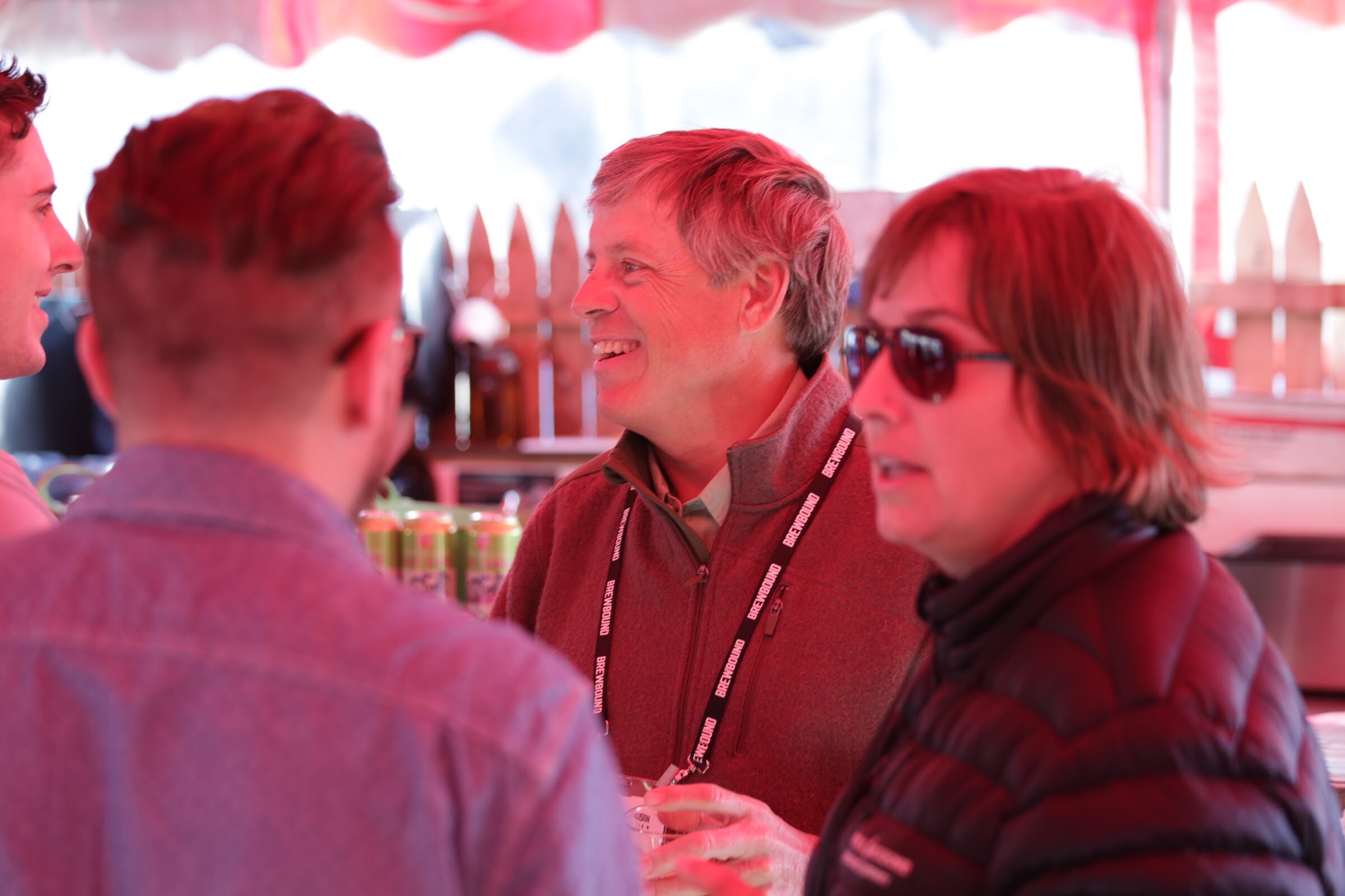 Group of attendees with lanyards talking and smiling in a tent lit red, with cans on the bar behind.