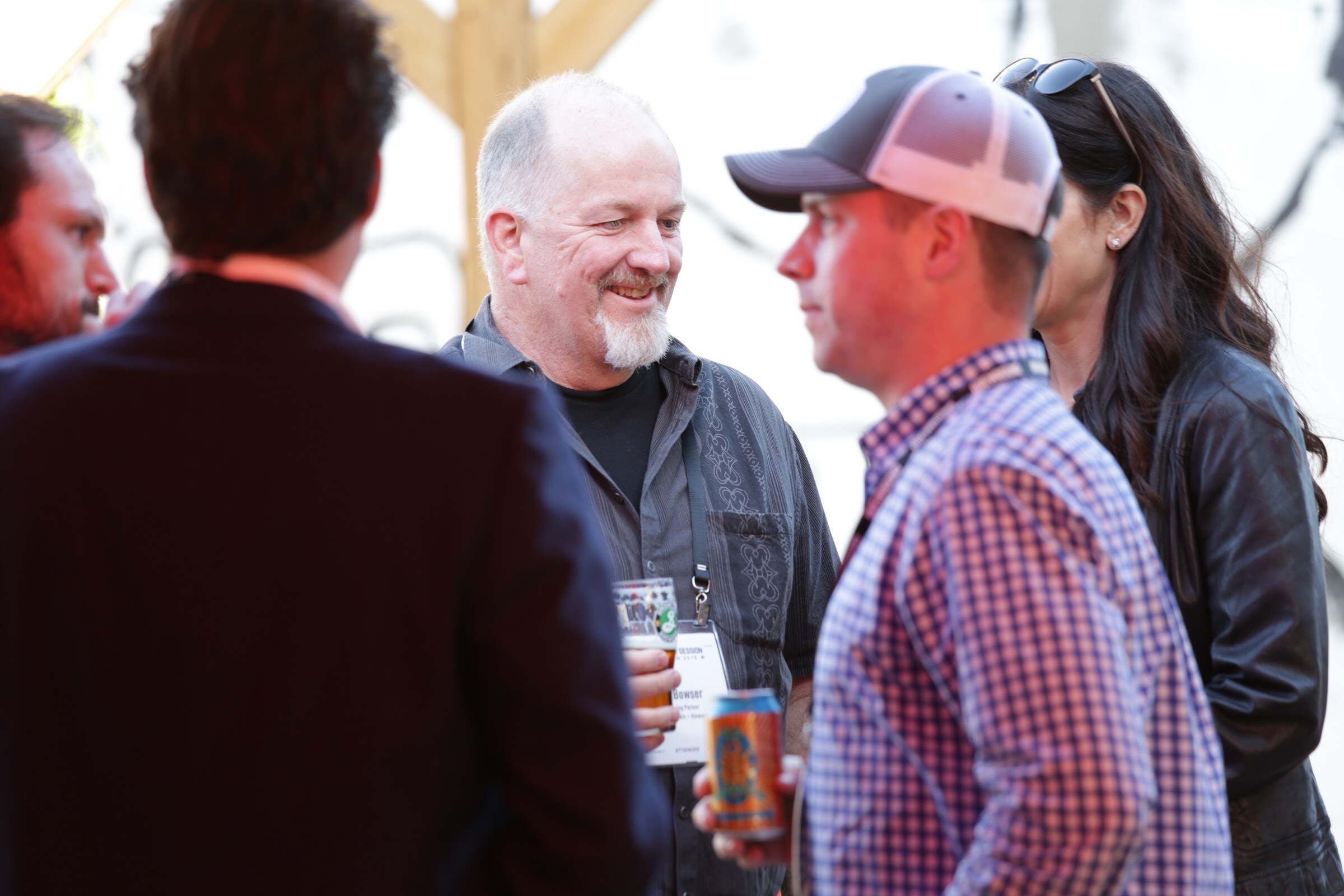 Group of attendees holding beer cups and cans and talking, one in a trucker cap, in bright light under a tent.