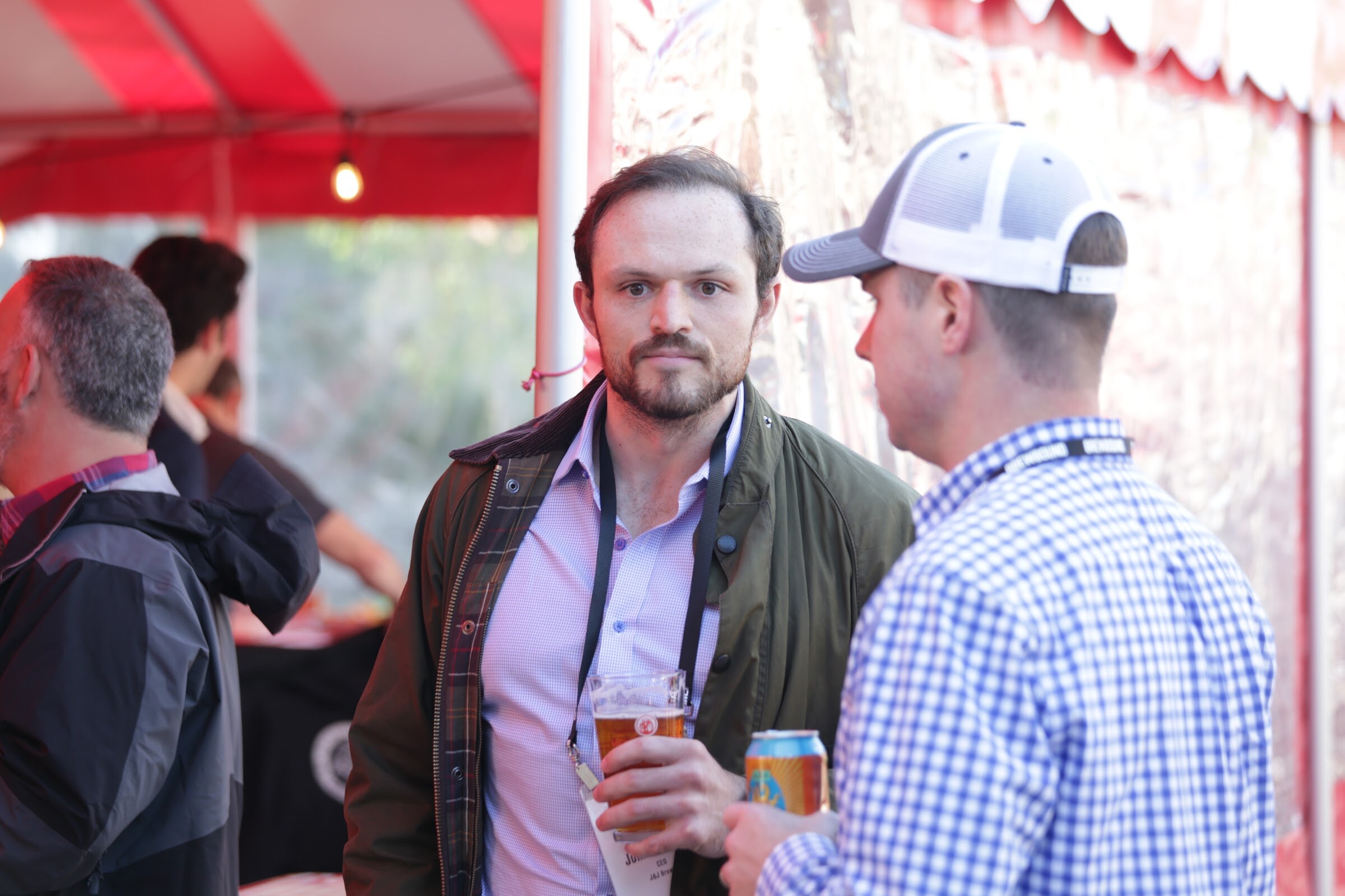 Man in an olive jacket holding a beer cup looking at the camera while another man in a trucker cap faces him, under a striped tent.