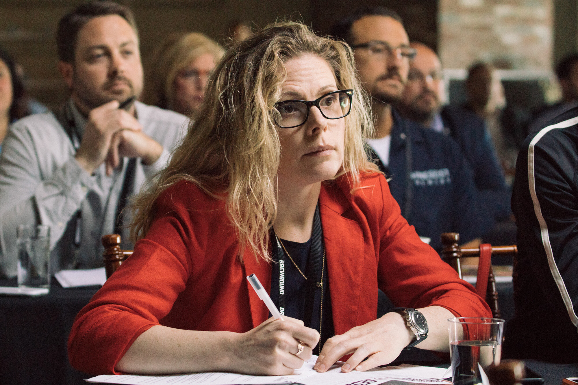 Woman with glasses and wavy hair in a red blazer holding a pen over paper, with other attendees seated behind.