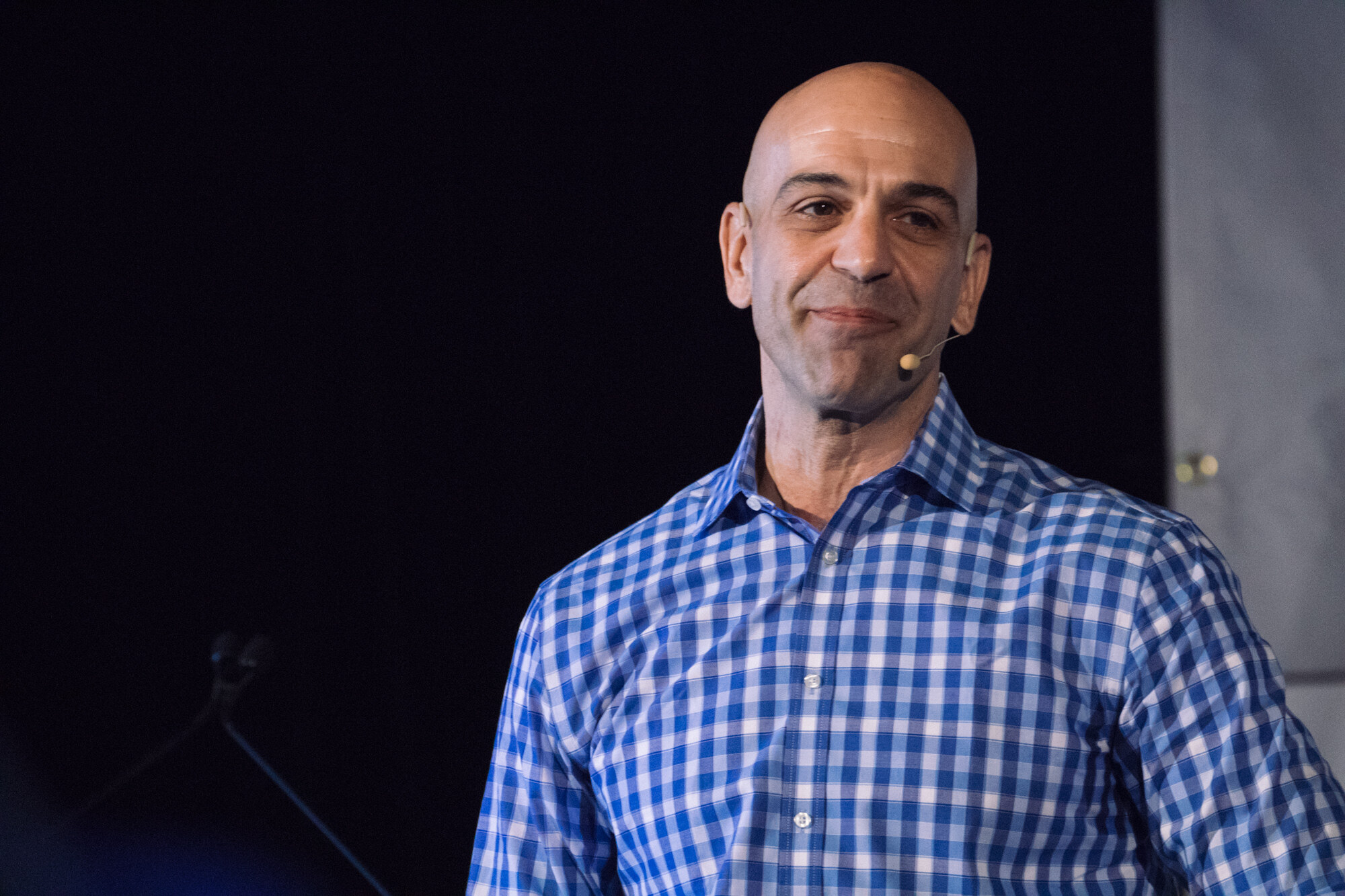 Bald man in a blue gingham shirt with a headset microphone smiling slightly against a dark background.