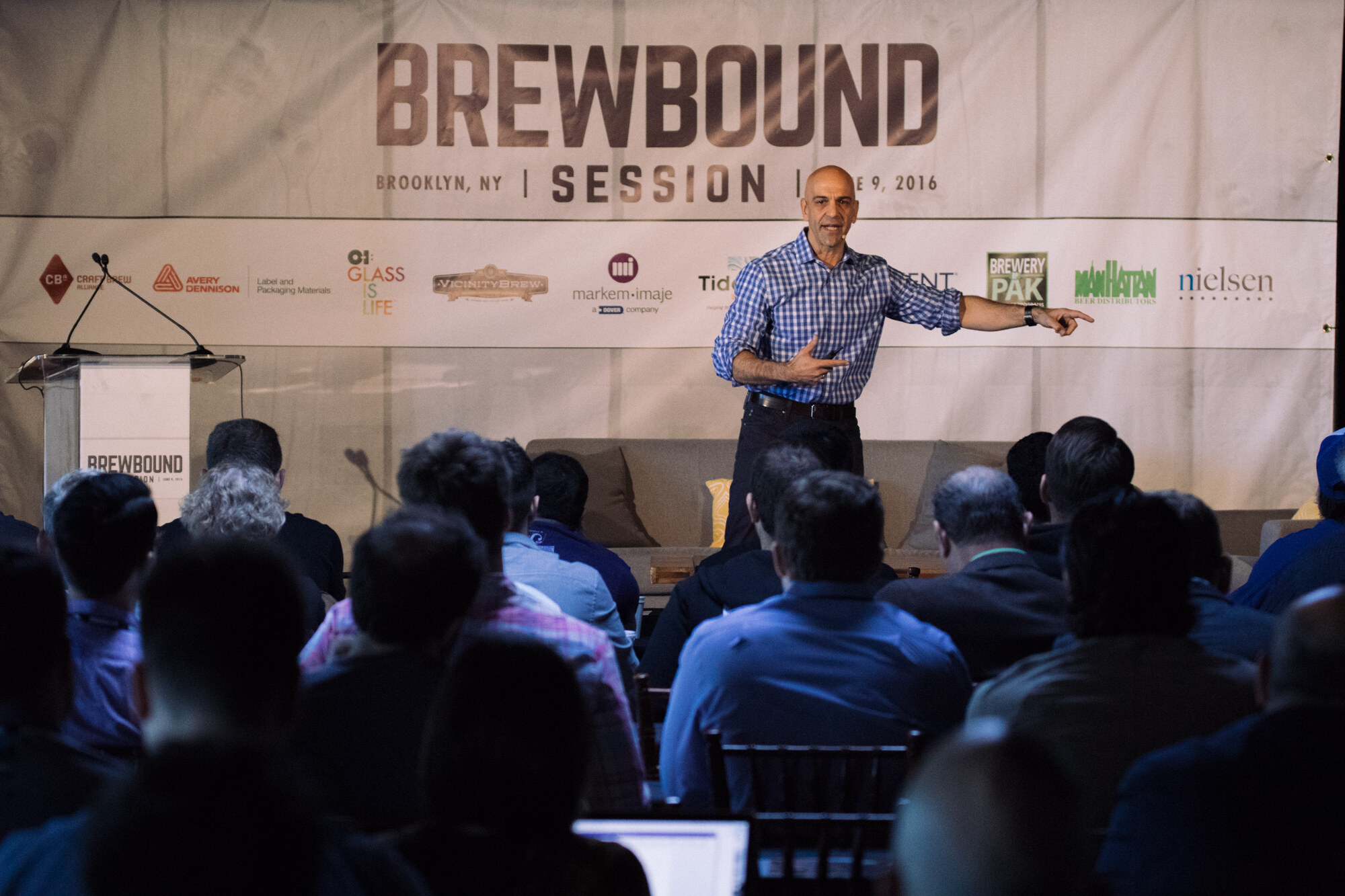 Man in a blue checked shirt pointing while speaking on stage in front of a Brewbound Session banner with sponsor logos, with the audience's heads in the foreground.