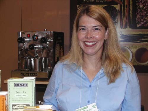 A smiling woman with long hair and a light blue shirt stands at a booth beside an espresso machine and boxes of Tazo tea, including Matcha.