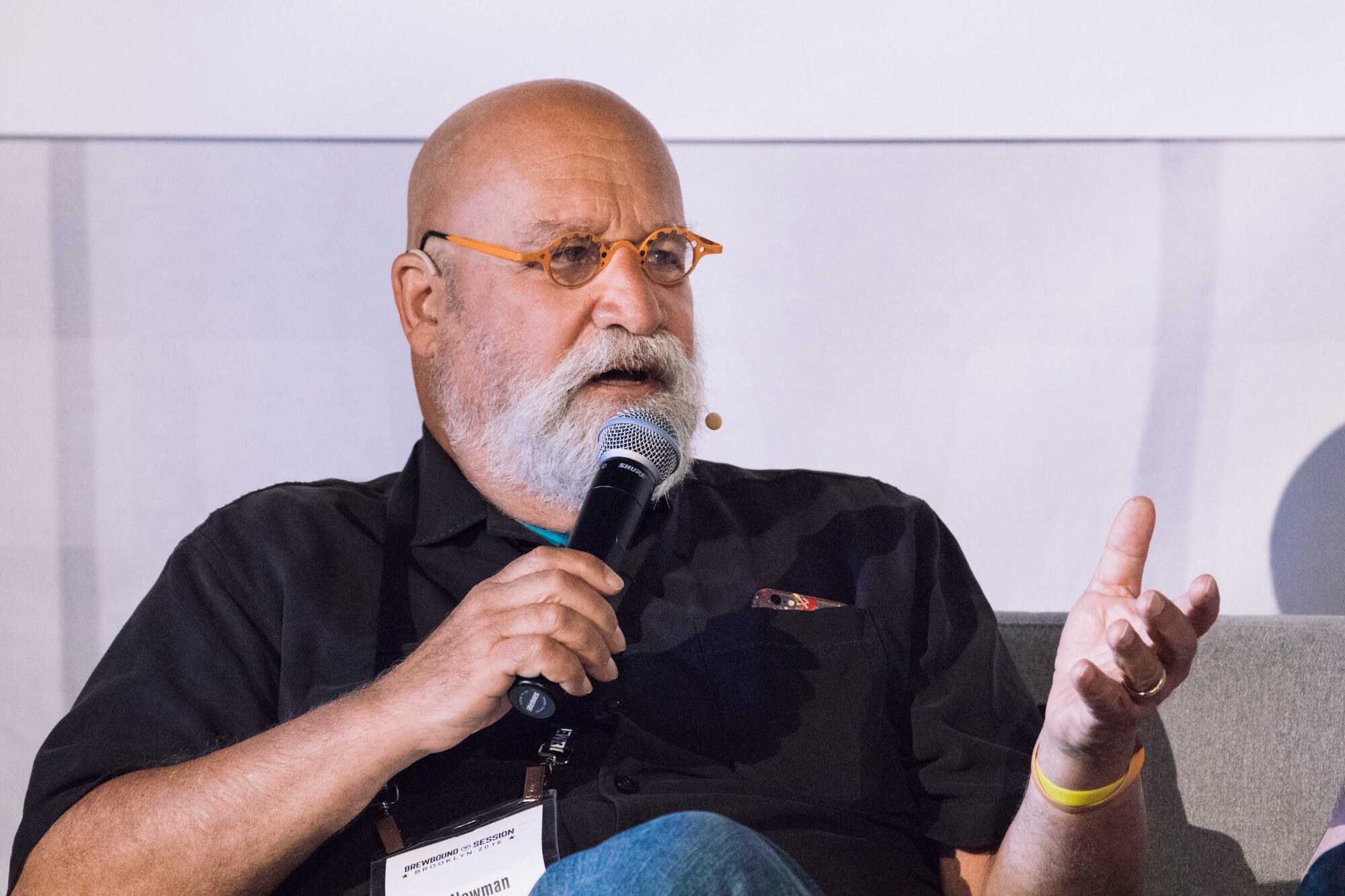 Bald man with a white beard and orange glasses holding a microphone and gesturing with one hand.