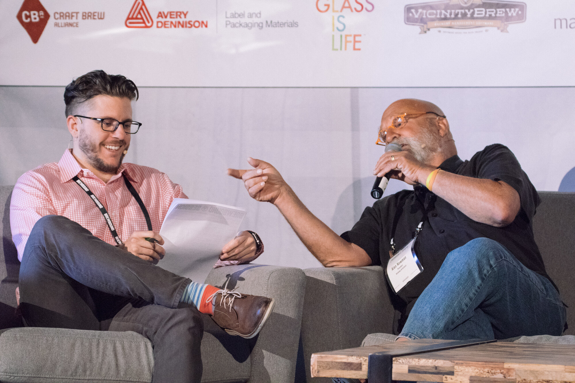 Two men seated on gray couches, one holding papers and smiling, the other pointing and holding a microphone, beneath Craft Brew Alliance and Avery Dennison sponsor logos.