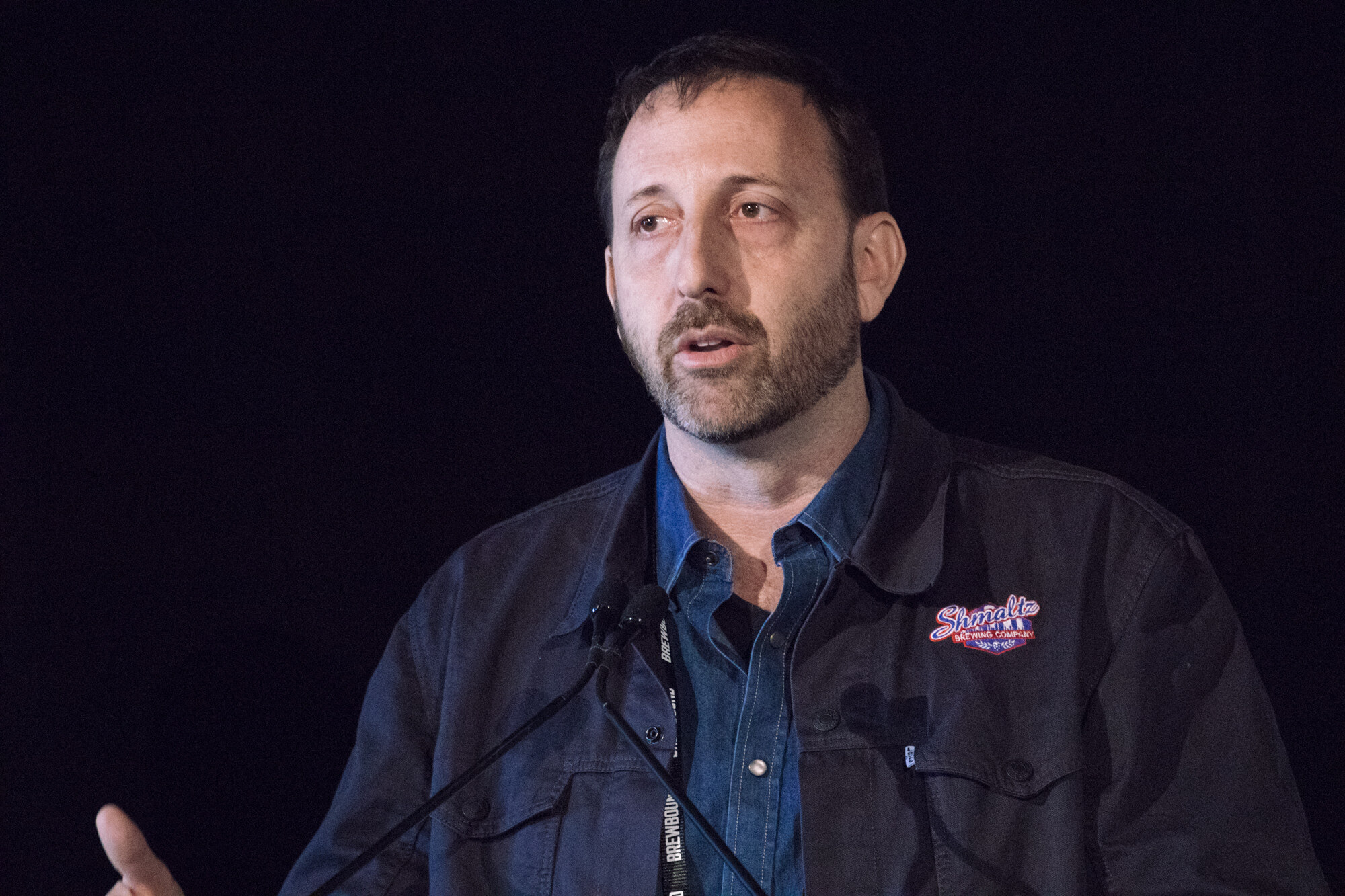 Bearded man in a dark jacket with a brewery logo patch speaking into a podium microphone.