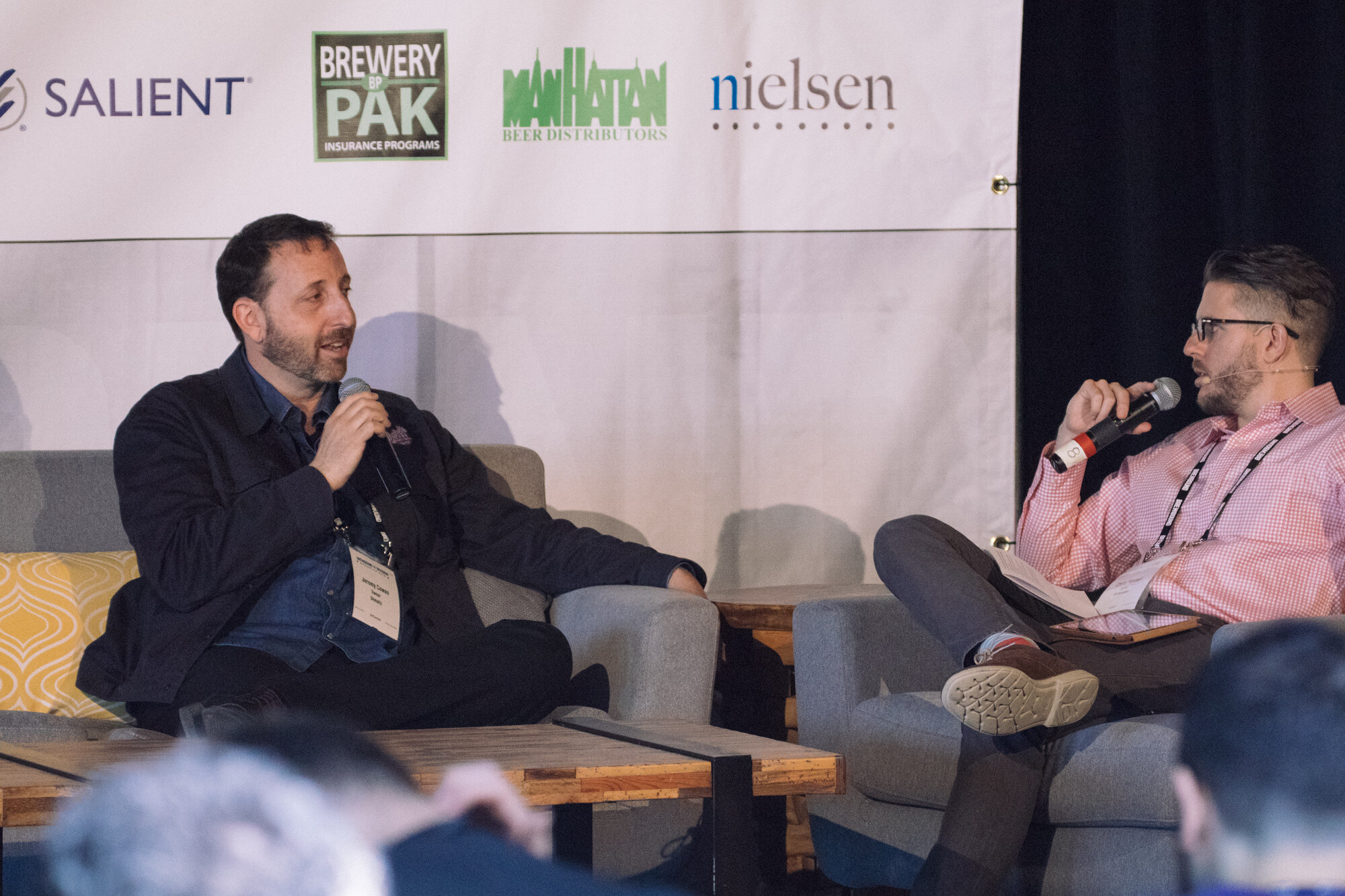 Two men seated in armchairs with microphones in front of a banner with Salient, Brewery Pak, Manhattan Beer Distributors and Nielsen logos.