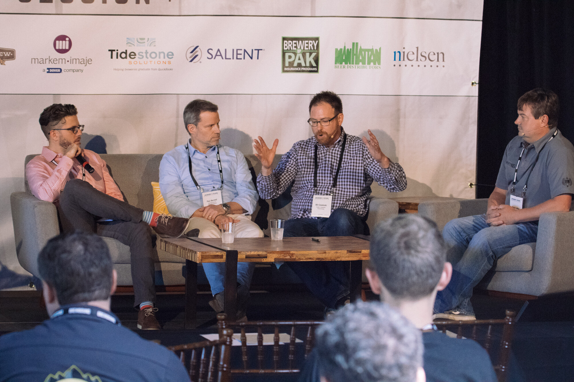 Four men on gray couches around a wood coffee table, one gesturing with both hands, under a sponsor banner.