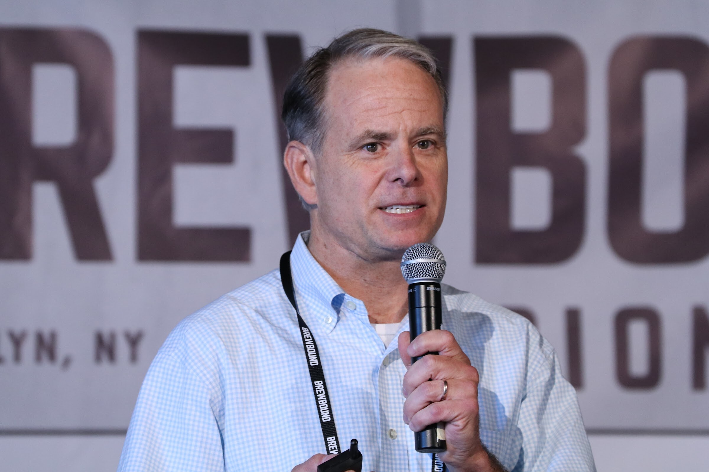 Man in a light blue checked shirt holding a microphone and presentation clicker in front of the Brewbound banner.