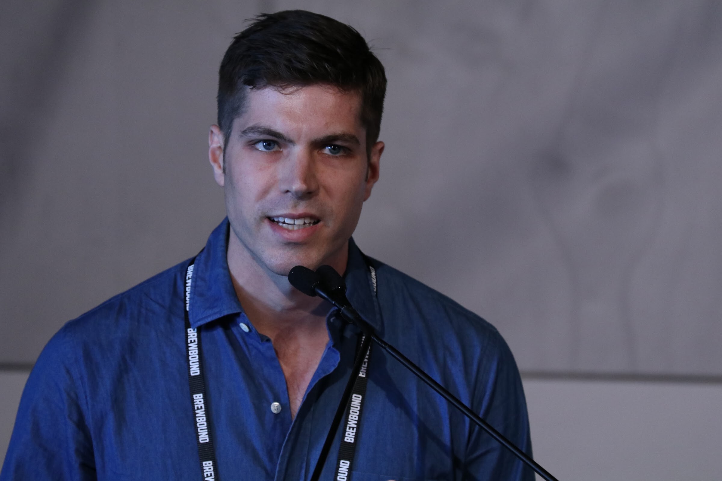 Dark-haired man in a blue shirt with a Brewbound lanyard speaking into a podium microphone.