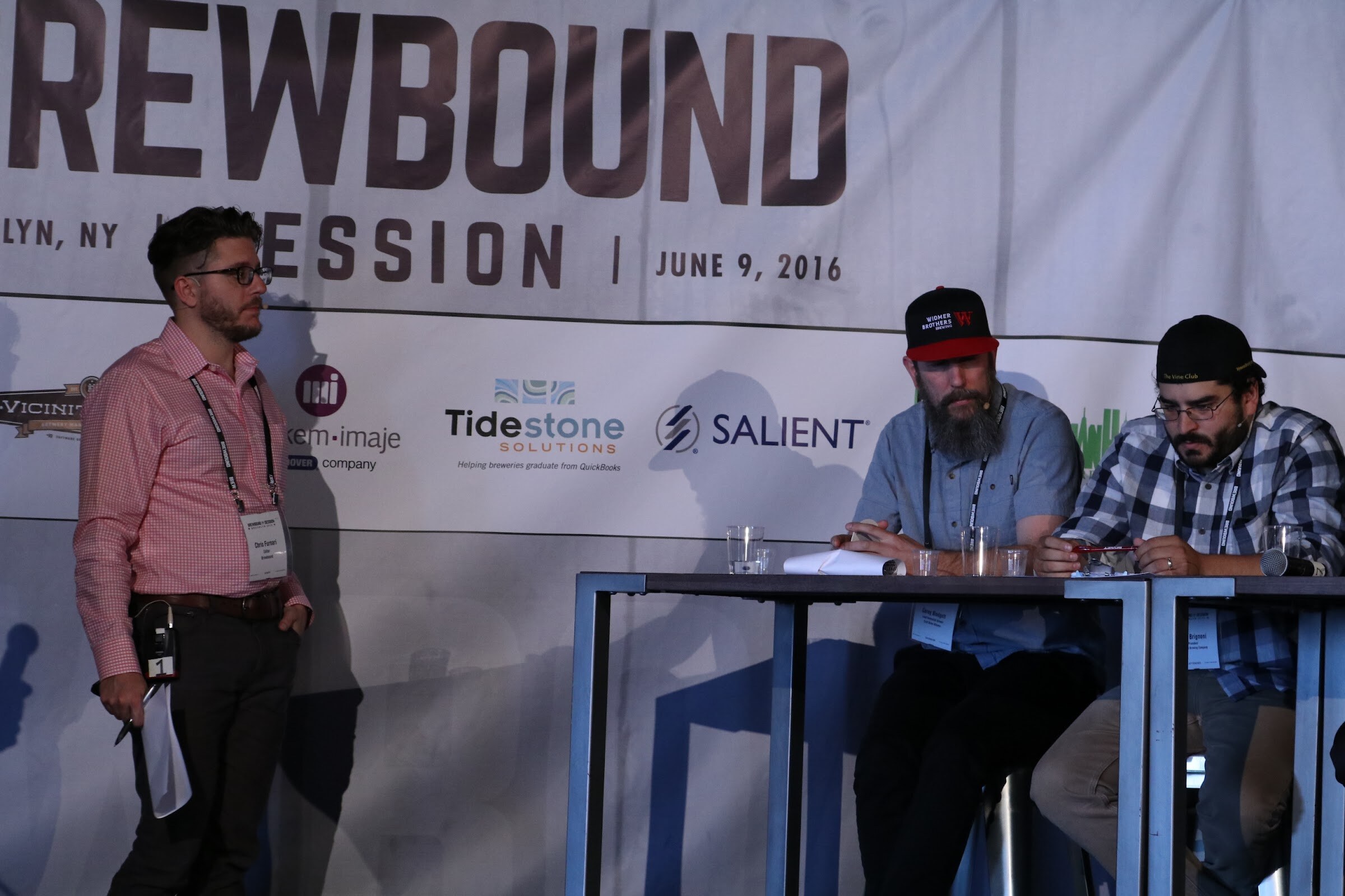 Man in a pink checked shirt standing beside two seated men at a high table under the Brewbound Session banner dated June 9, 2016.