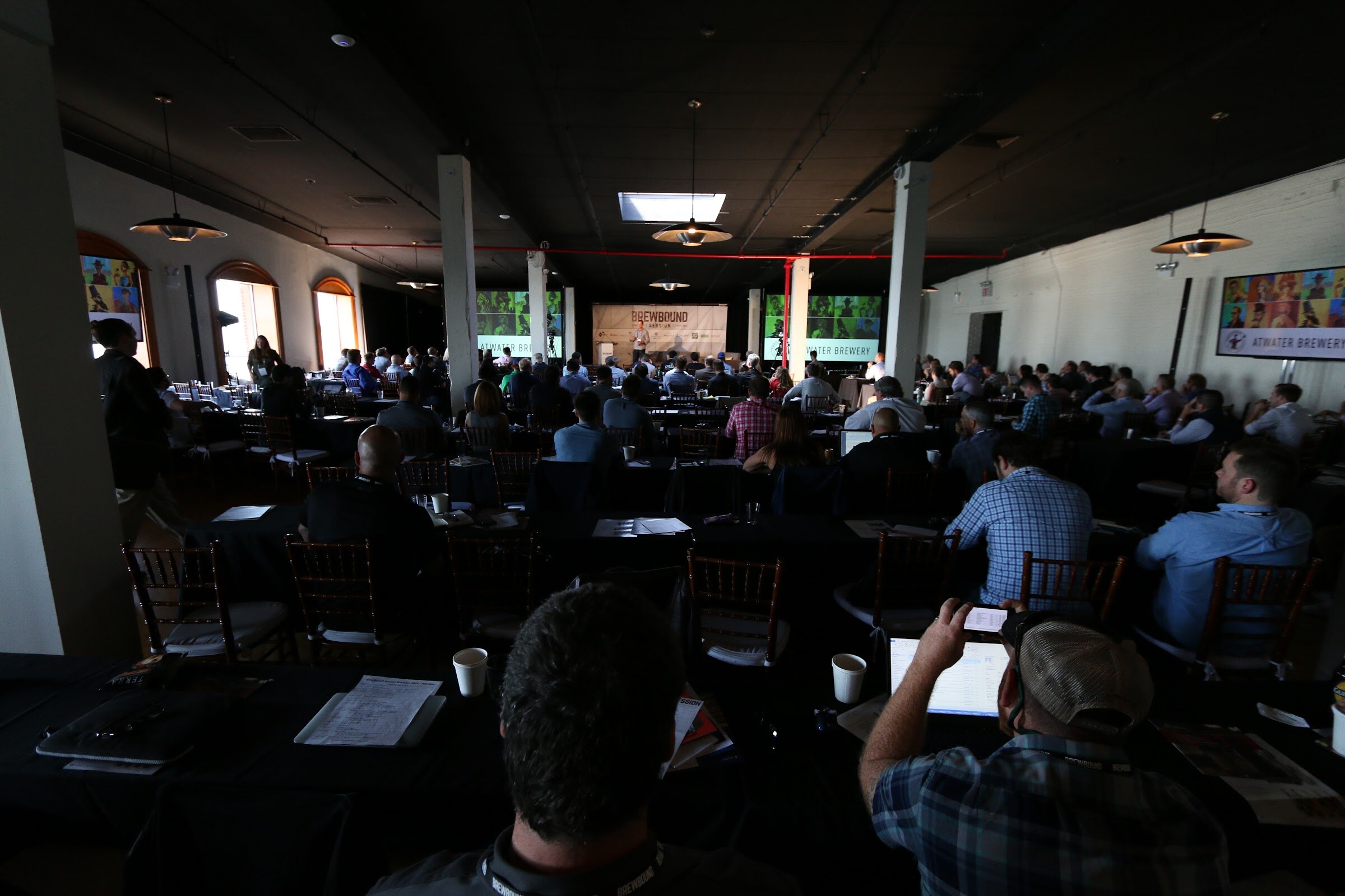 Wide view of a darkened event hall with attendees at tables facing a stage with a Brewbound banner and screens.
