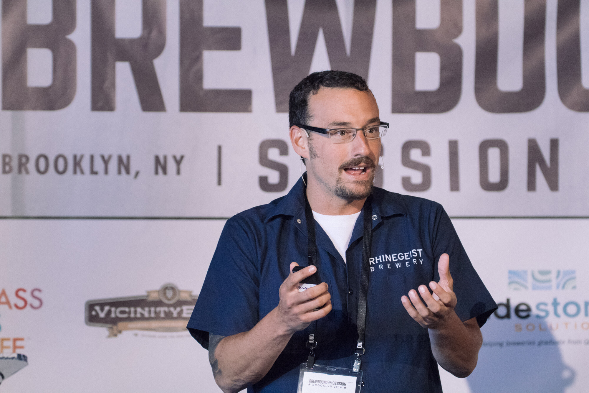 Man in a navy Rhinegeist Brewery work shirt with a lanyard speaking in front of a Brewbound Session Brooklyn banner.