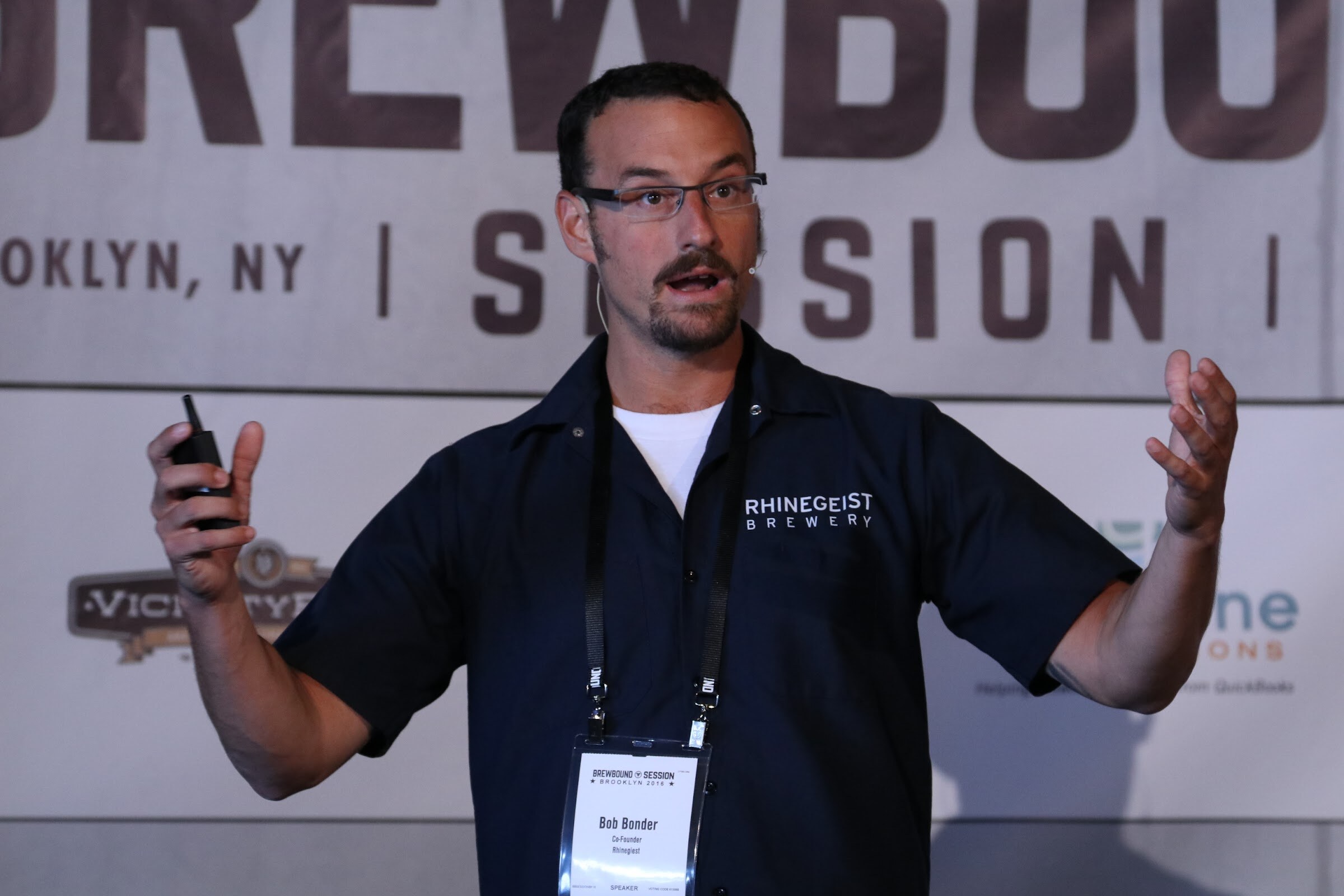 Man in a navy Rhinegeist Brewery shirt holding a clicker with arms outstretched in front of a Brewbound Session banner.