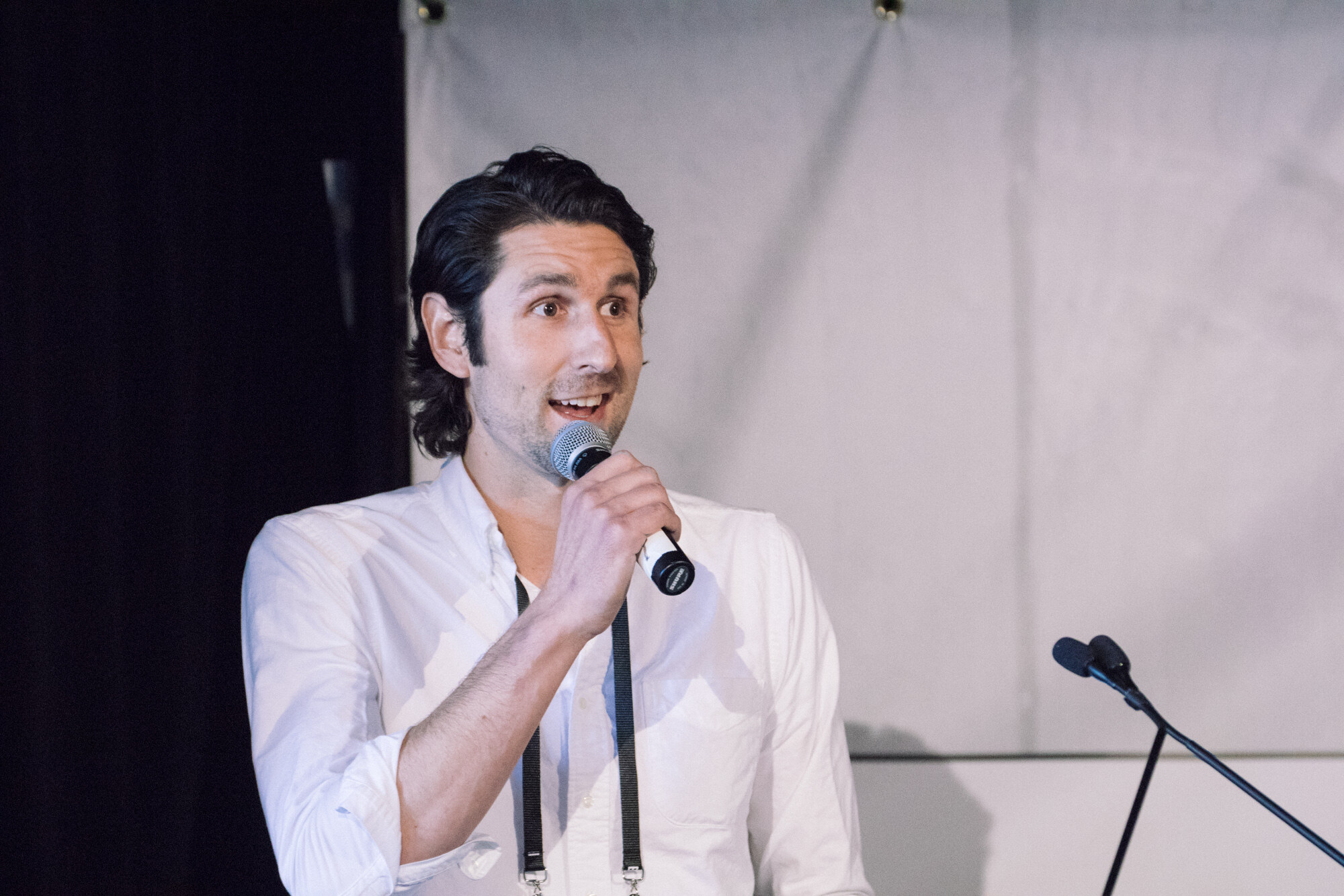 Man with dark wavy hair in a white shirt speaking into a handheld microphone beside a podium.