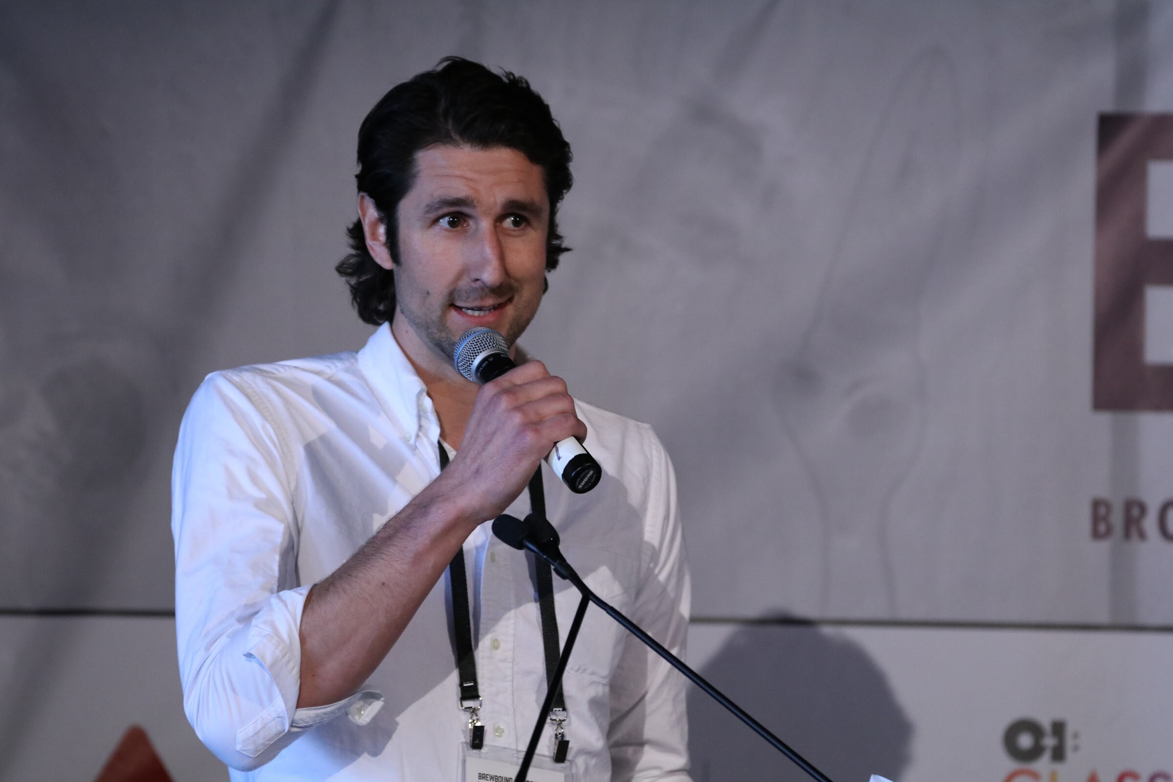 Man with dark wavy hair in a white open-collar shirt holding a microphone at a lectern.